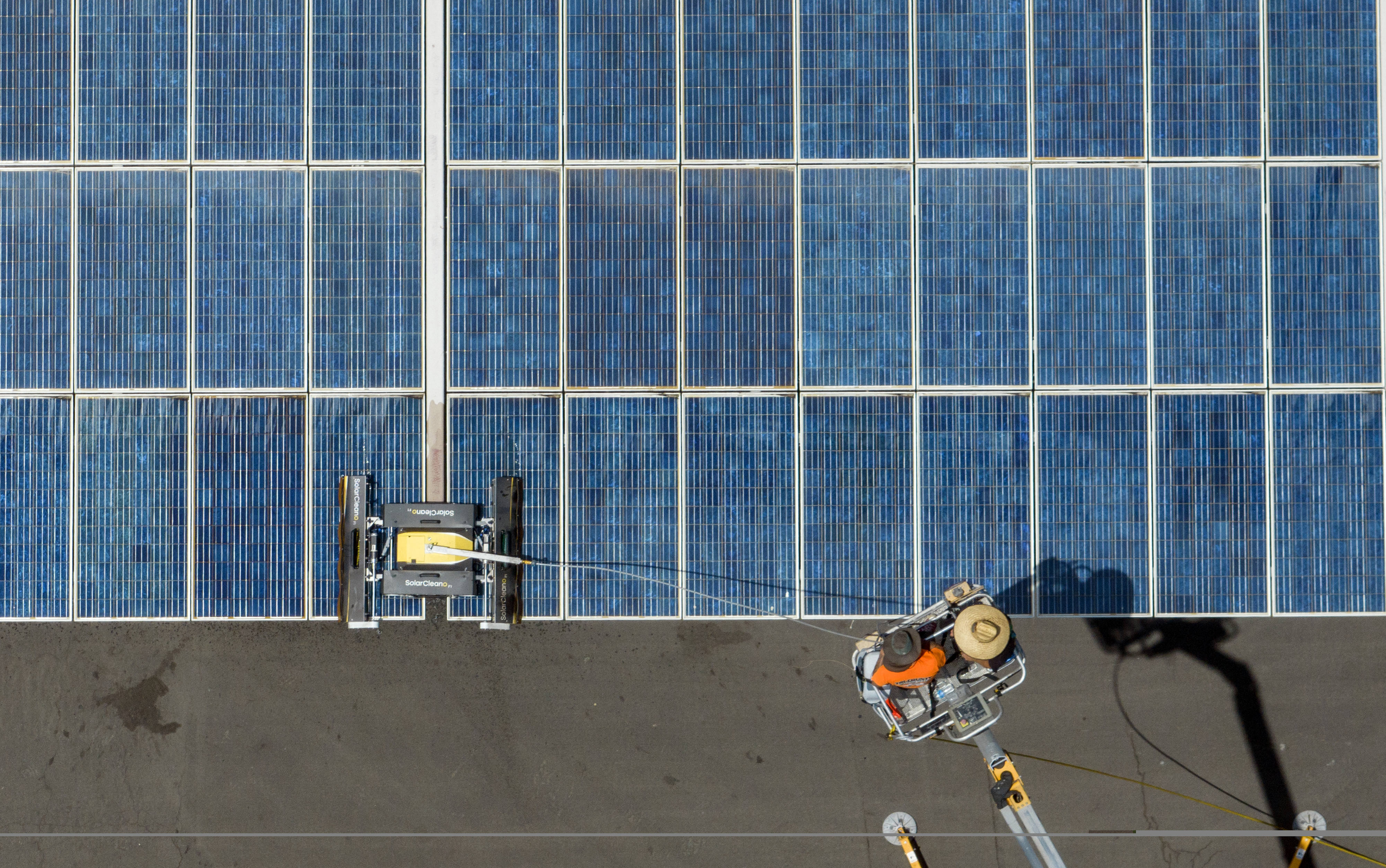 A person wearing a straw hat and riding in the bucket of an aerial lift is seen from above as they control a robot washing solar panels.