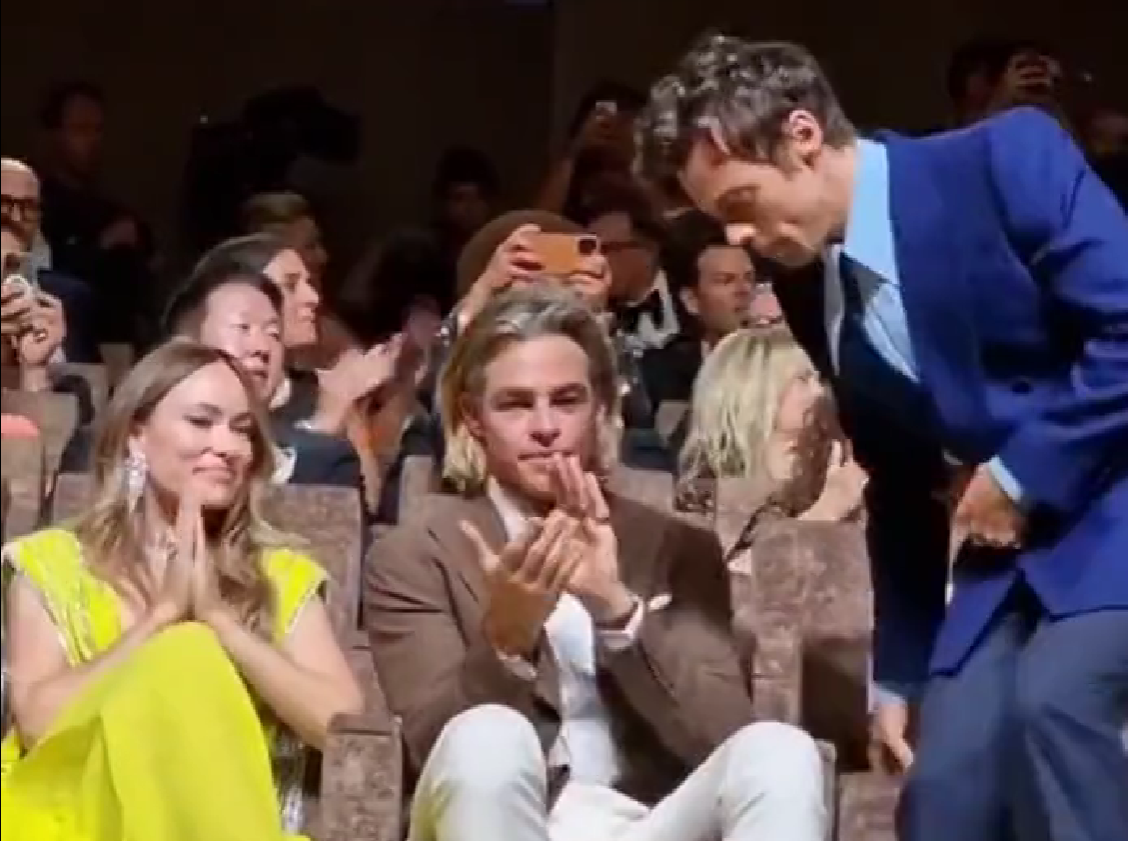 At a galmorous premiere screening, Harry Styles, in a blue suit, leans over a seated Chris Pine, who is applauding. It looks like Styles might be spitting.