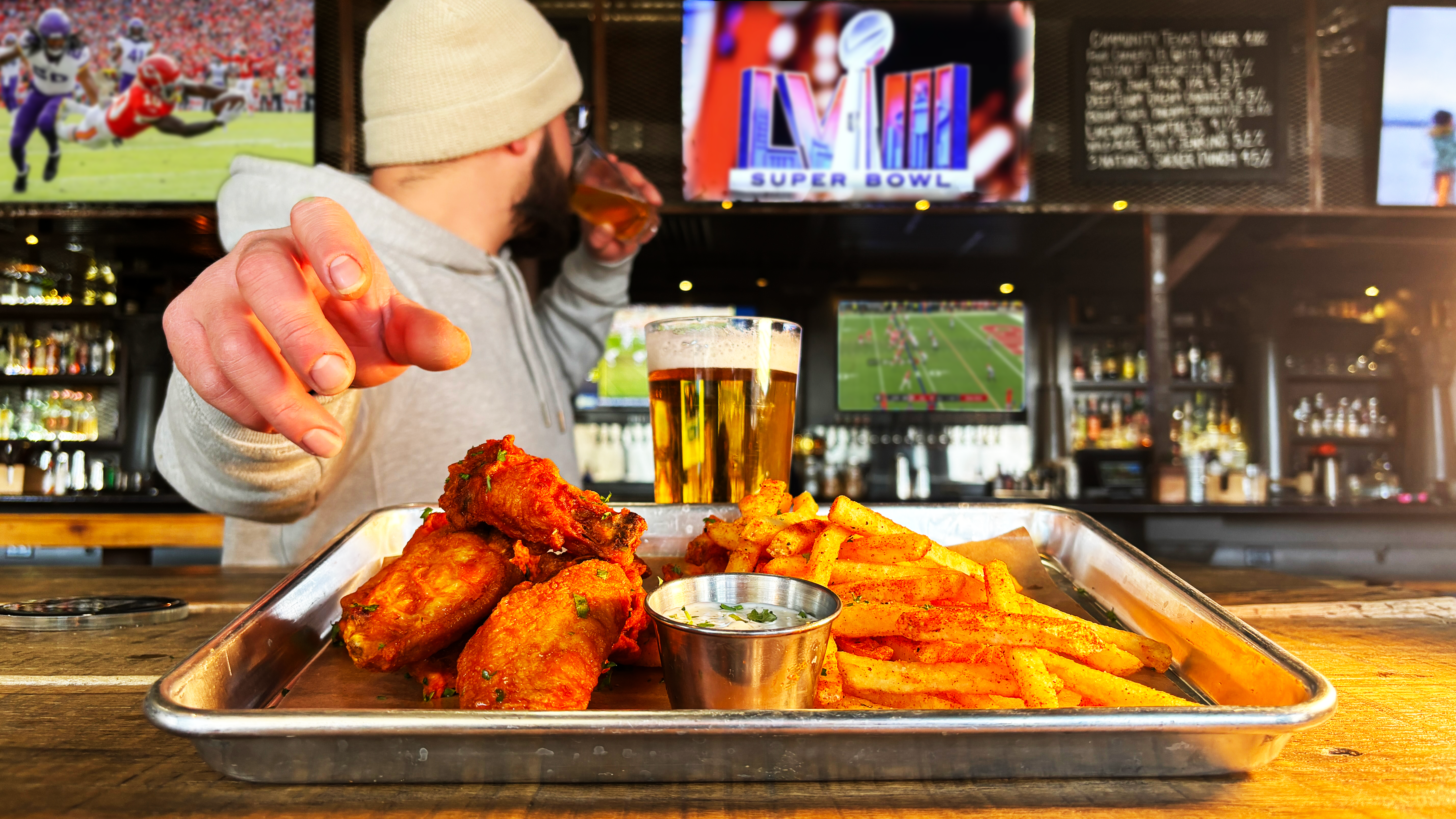 A man drinks a beer with his head turned towards a TV behind him. His hand reaches for a plate of wings and fries in front of the camera. 