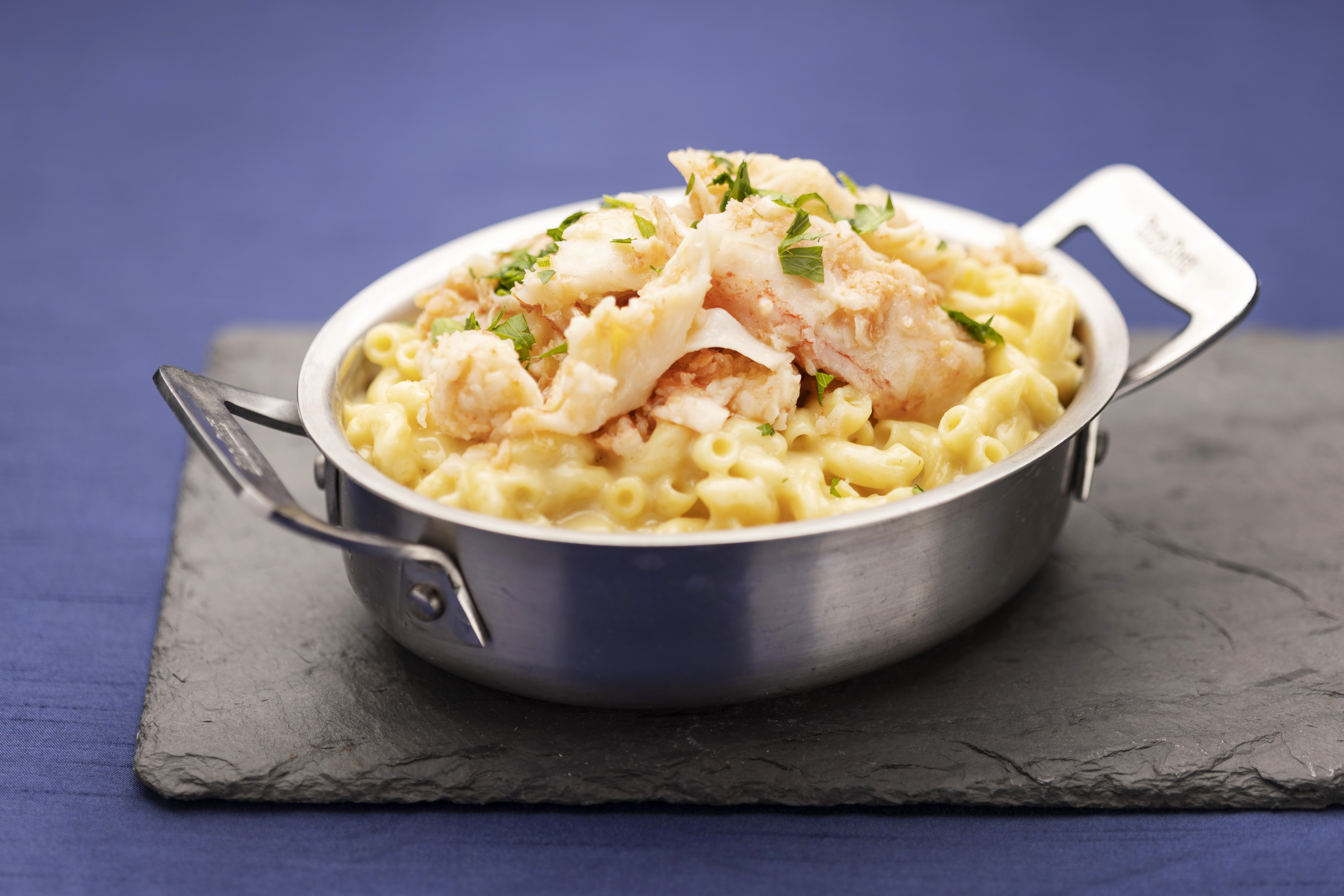 In a silver serving pot, mac and cheese is topped with chunks of lobster and herbs.