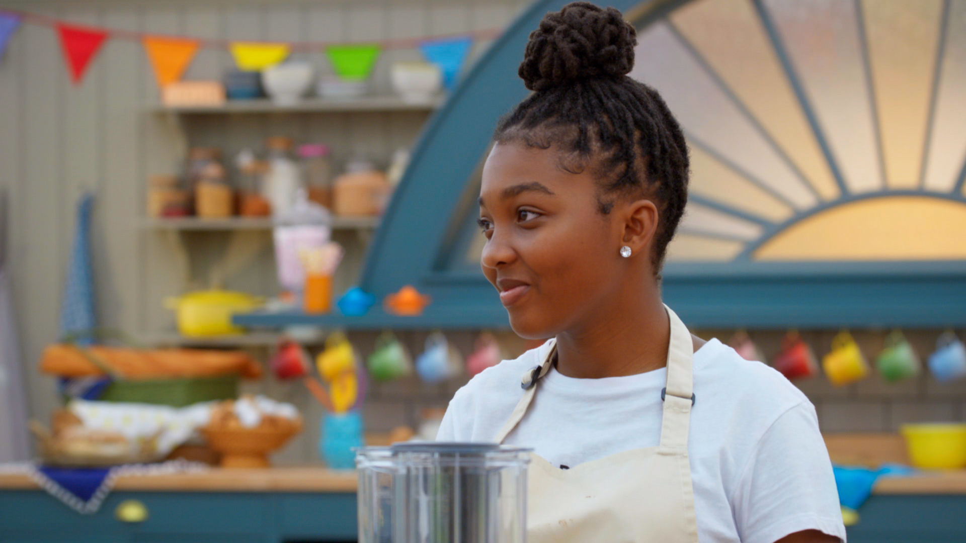 A young Black girl stands smiling in front of a blurred background of colorful kitchen appliances 
