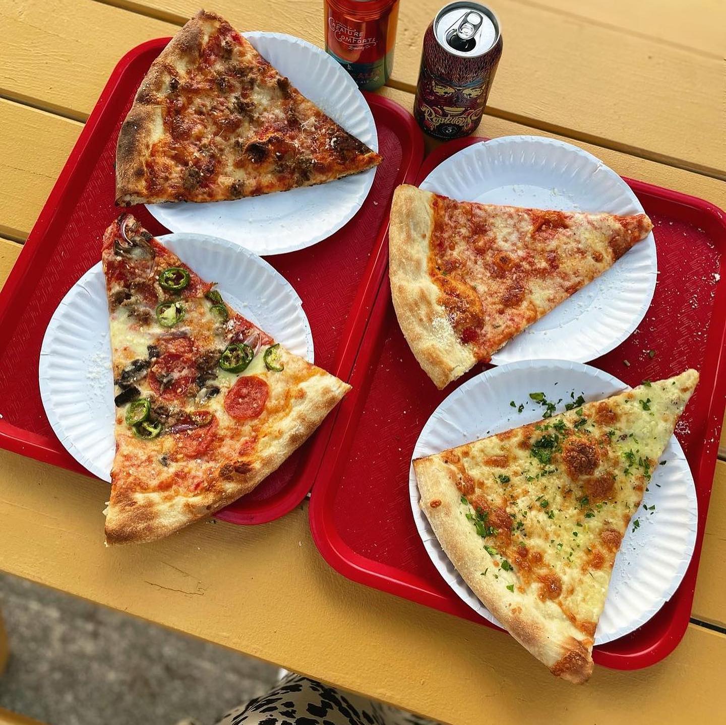 Four slices of pizza (sausage and onion, pepperoni and jalapenos, cheese, and garlic) sit on white paper plates on a red tray beside a two cans of beer.