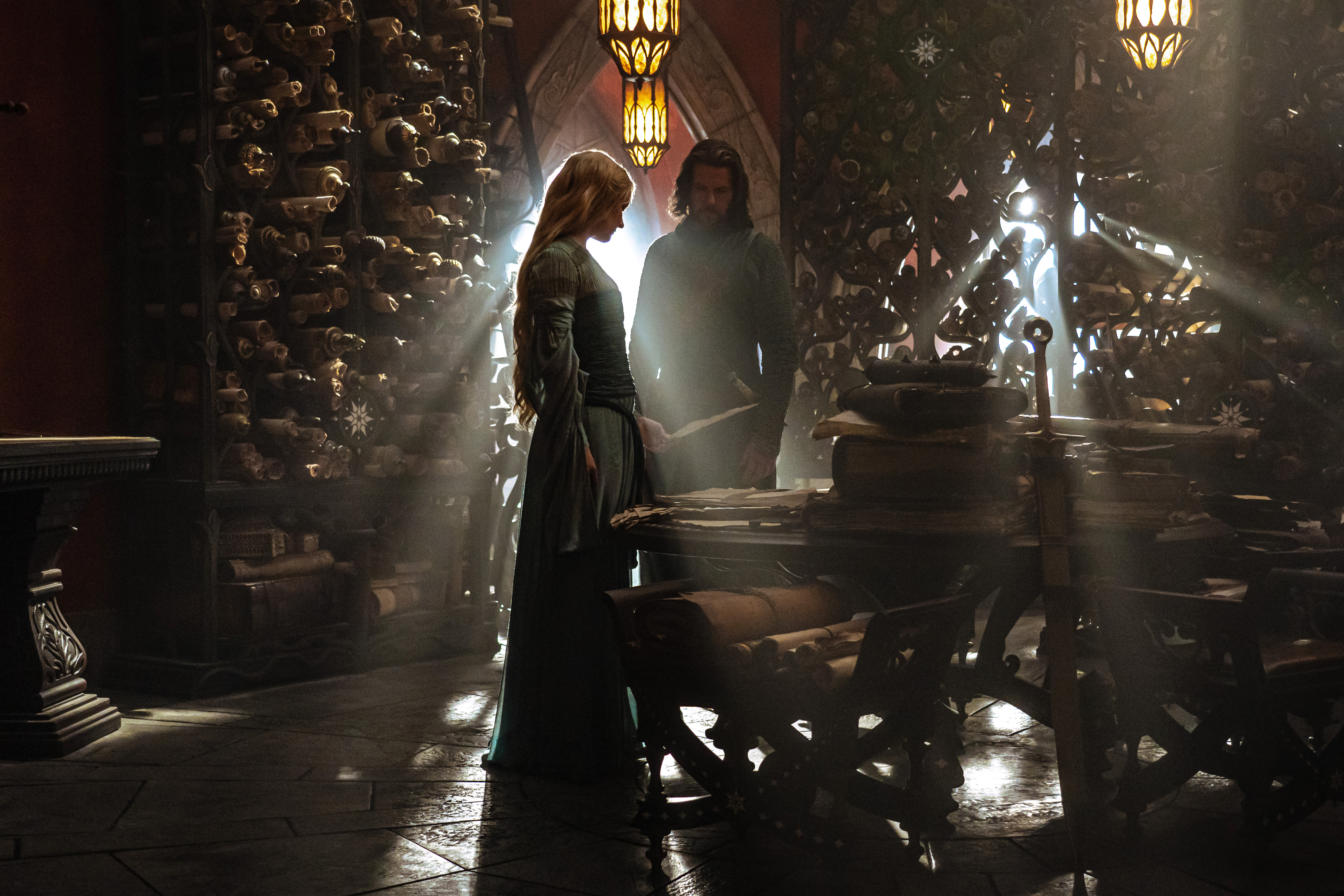 Galadriel and Elendil standing in a hall of records; both are silhouetted by a window with sunbeams around them, and looking at a scroll on the table