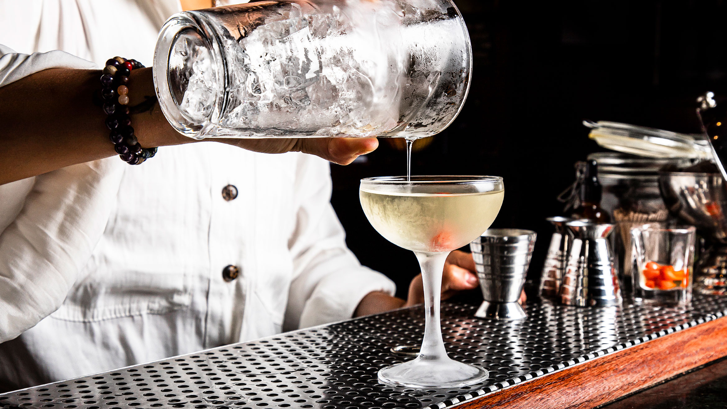 Figure behind a bar pours a liquid from a glass shaker into a coupe glass.