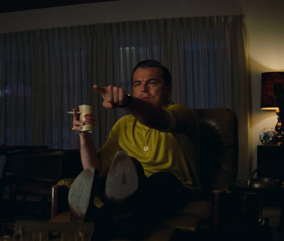 A still of Leonardo DiCaprio pointing in Once Upon a Time in Hollywood. He is sitting in a chair holding a beer and cigarette in one hand and pointing at the screen excitedly with the other