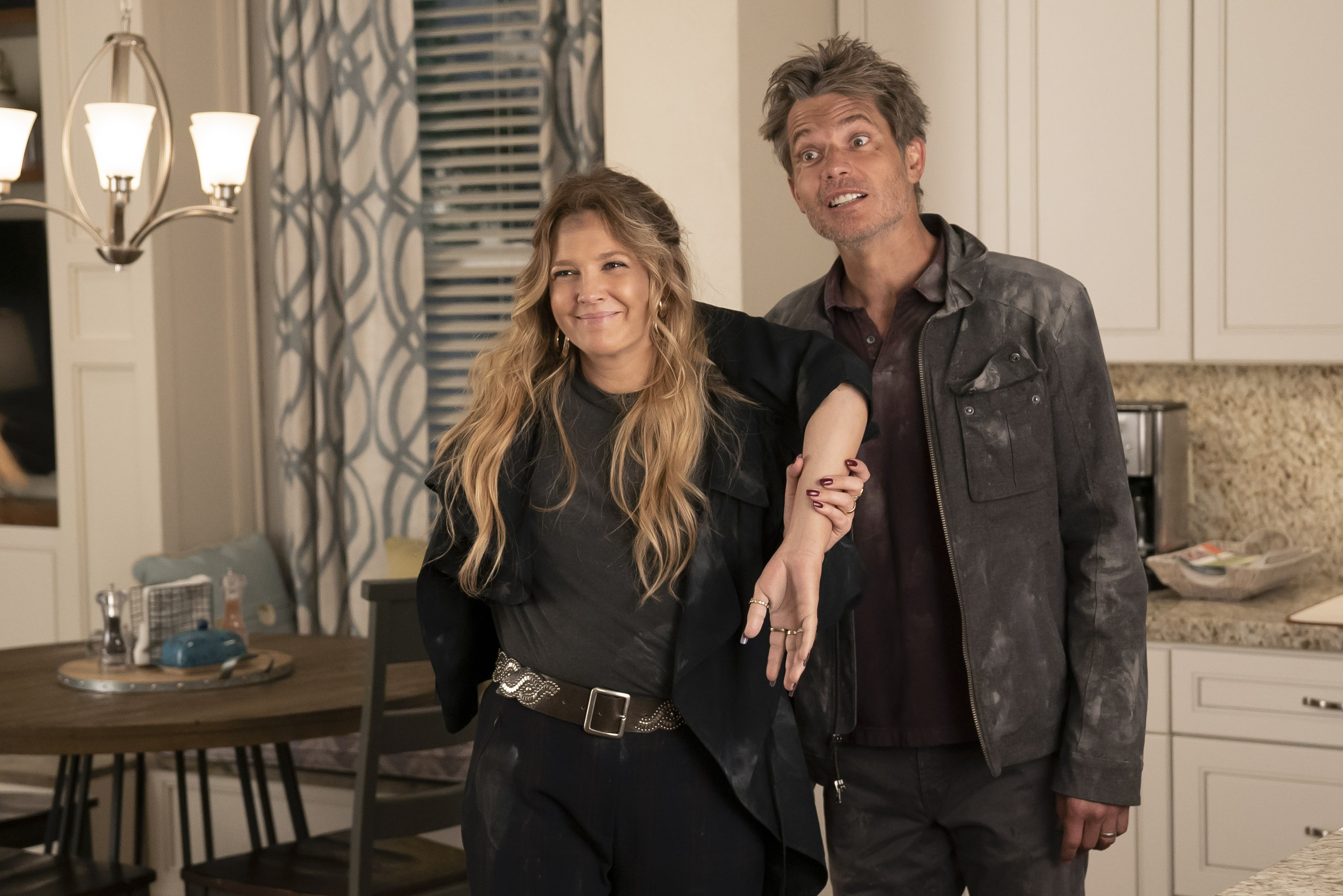 Drew Barrymore and Timothy Olyphant smile while covered in dirt and holding what appears to be a severed arm in Santa Clarita Diet.