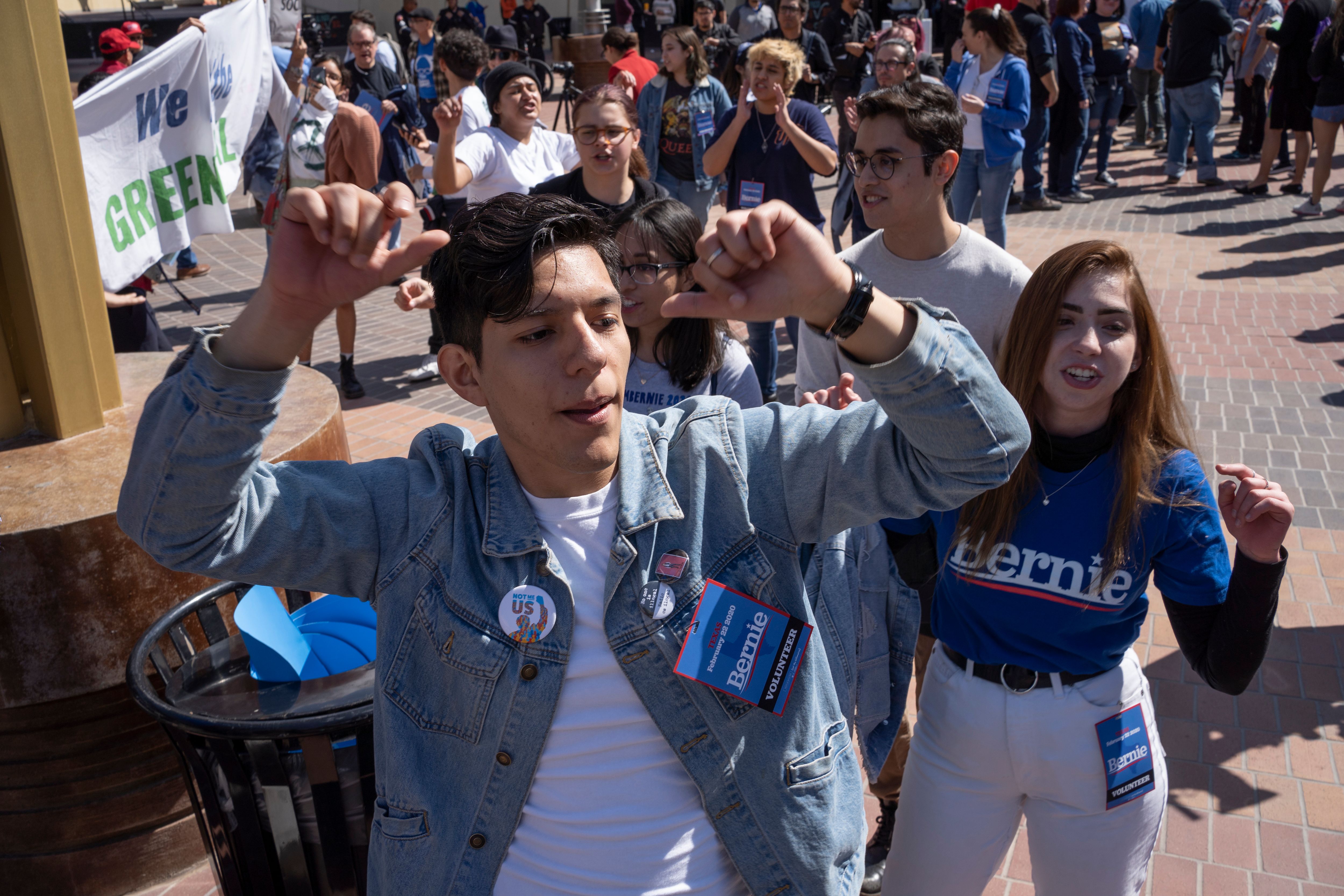 A young Latino man dances with his arms above his head in a blue jean jacket bearing a Bernie Sanders badge; a predominately young, Latino crowd dances in the sunshine behind him.