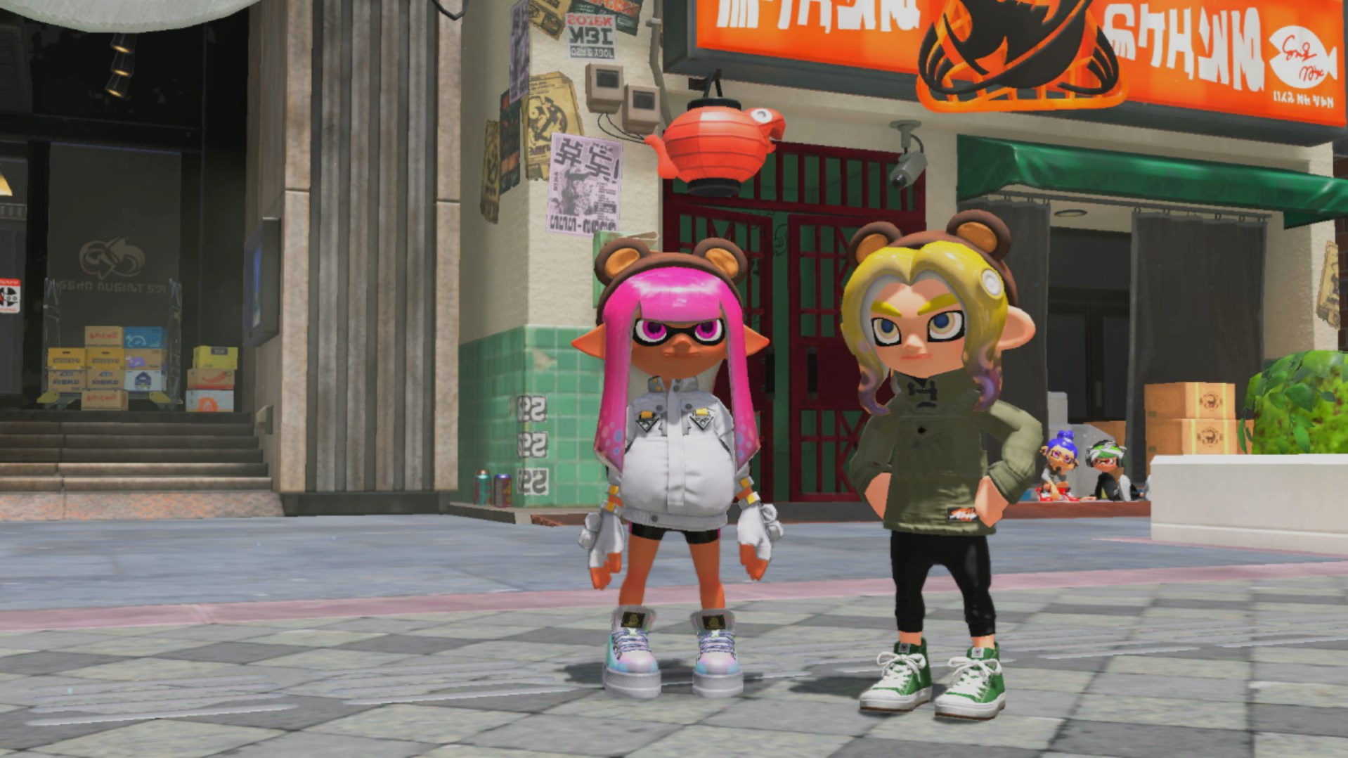 A pink inkling and yellow octoling stand in Inkopolis both wearing Teddy Band bear ears