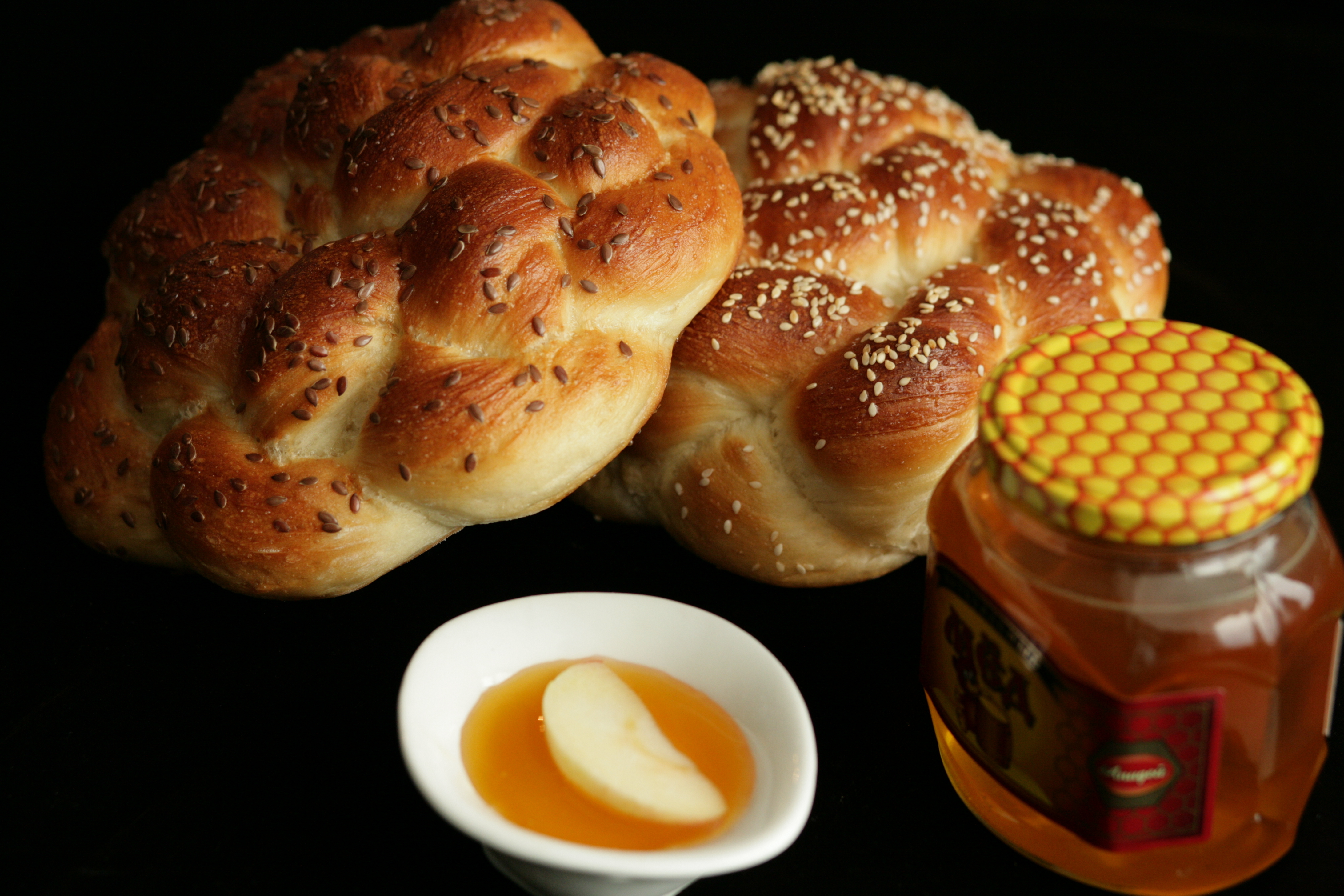 Two round braided challah next to a bowl of honey with a single apple slice in it and a jar of honey, on a black background. 