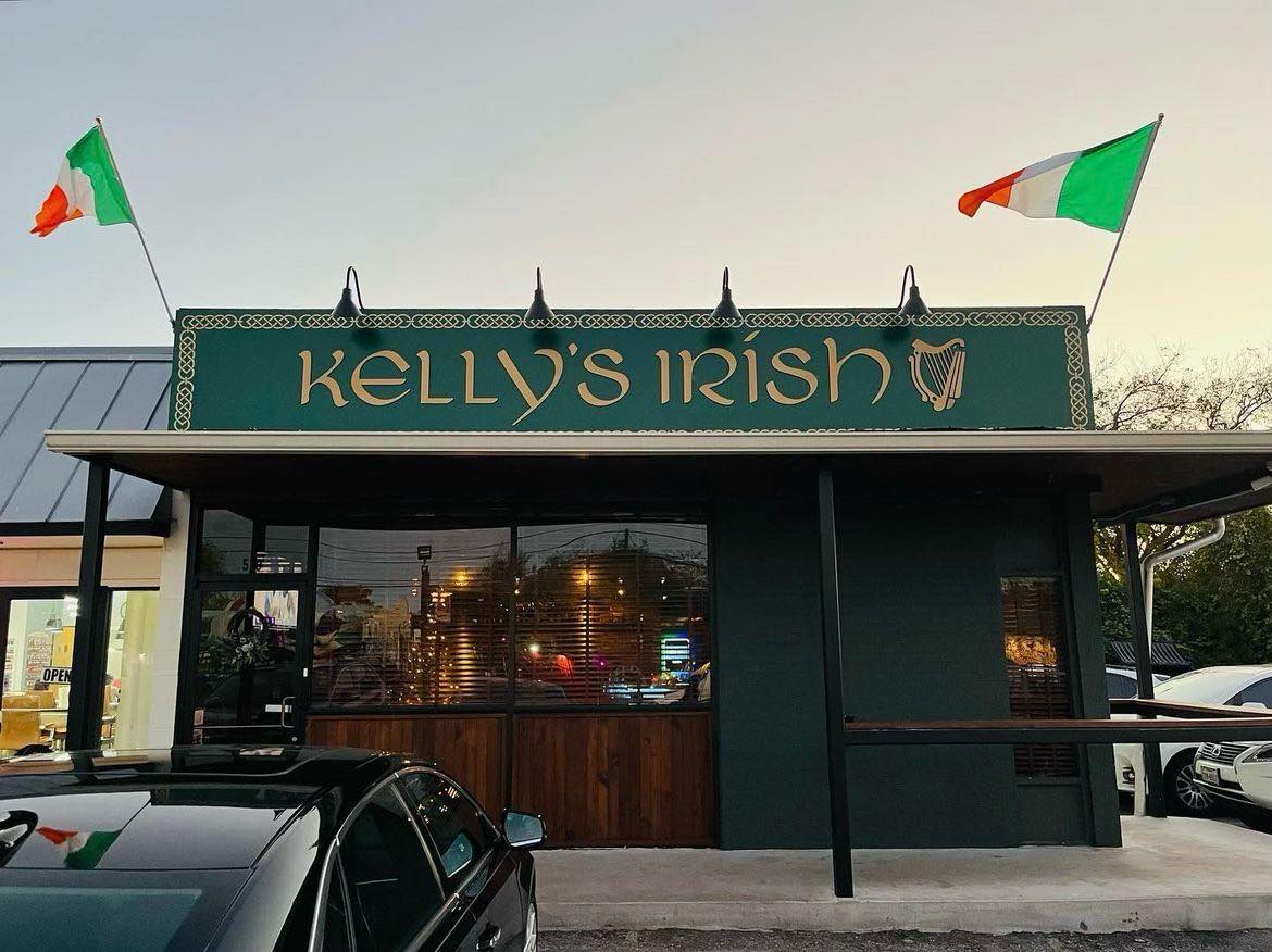 A bar facade with a green sign reading Kelly’s Irish flanked by Irish flags.