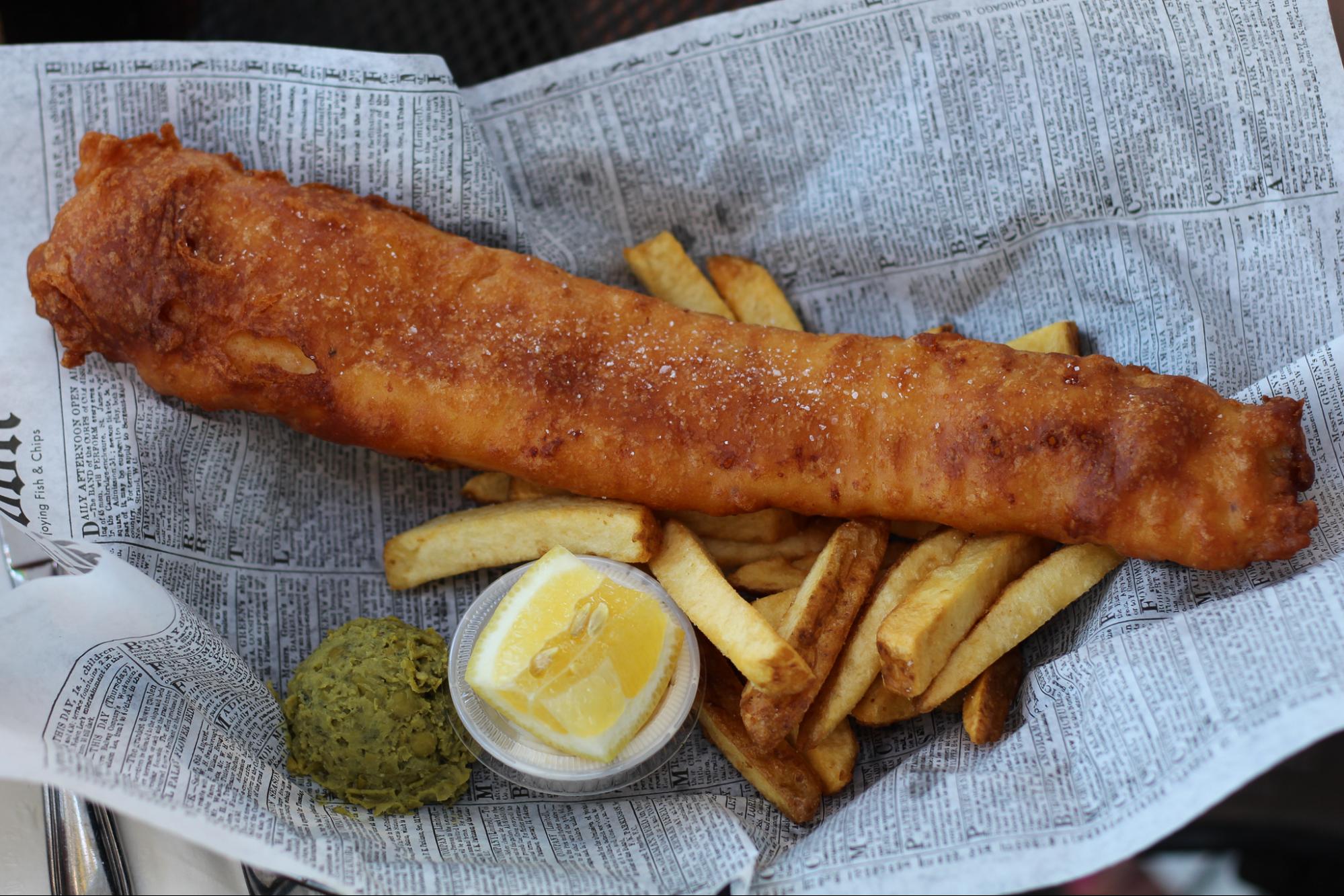 A 12-inch piece of fried cod with thick-cut fries, tartar sauce, and mushy pieces, served on a piece of newspaper. 