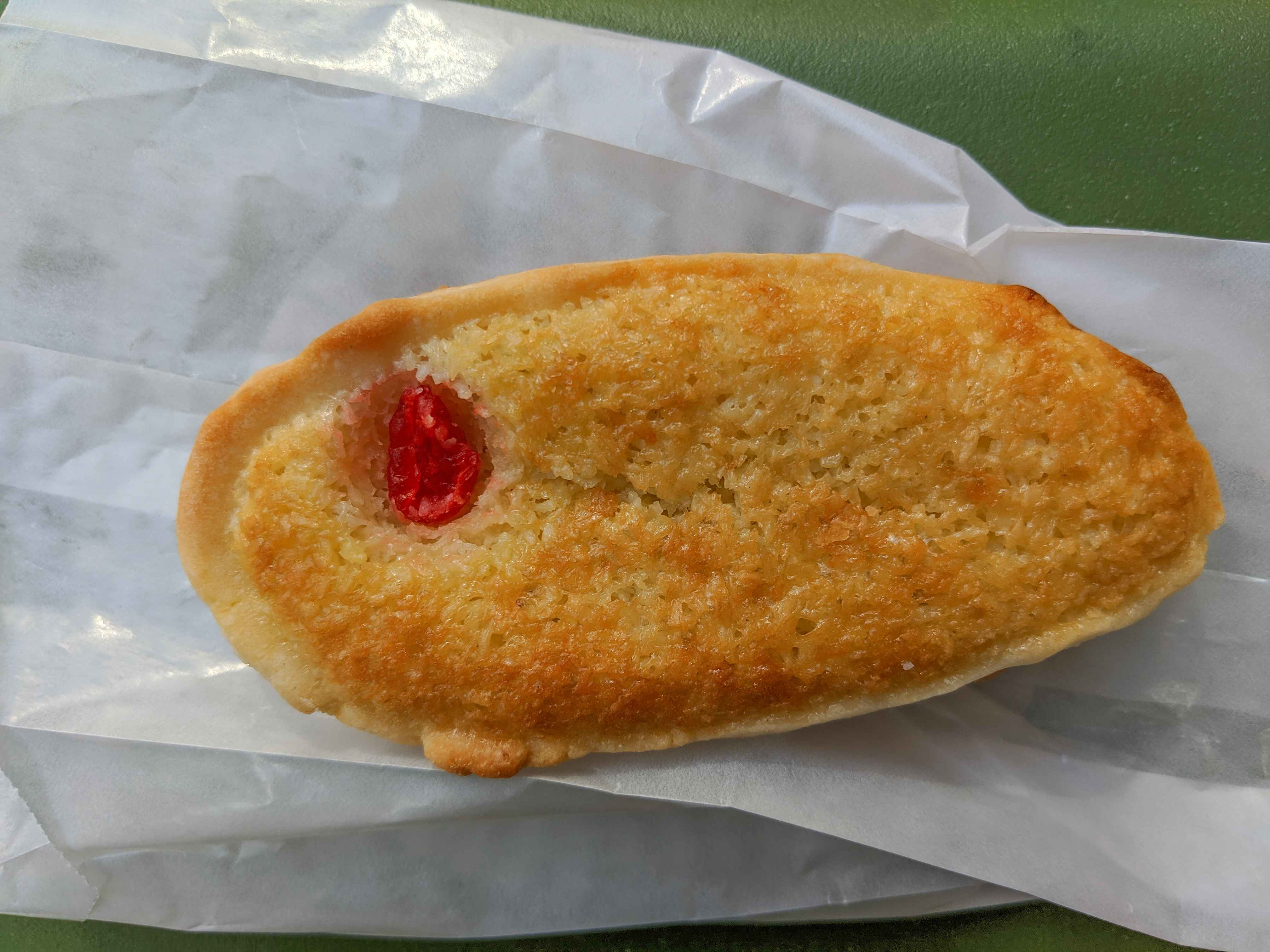 Overhead view of an oblong coconut tart with a maraschino cherry embedded in it, on a white paper bag on a green background