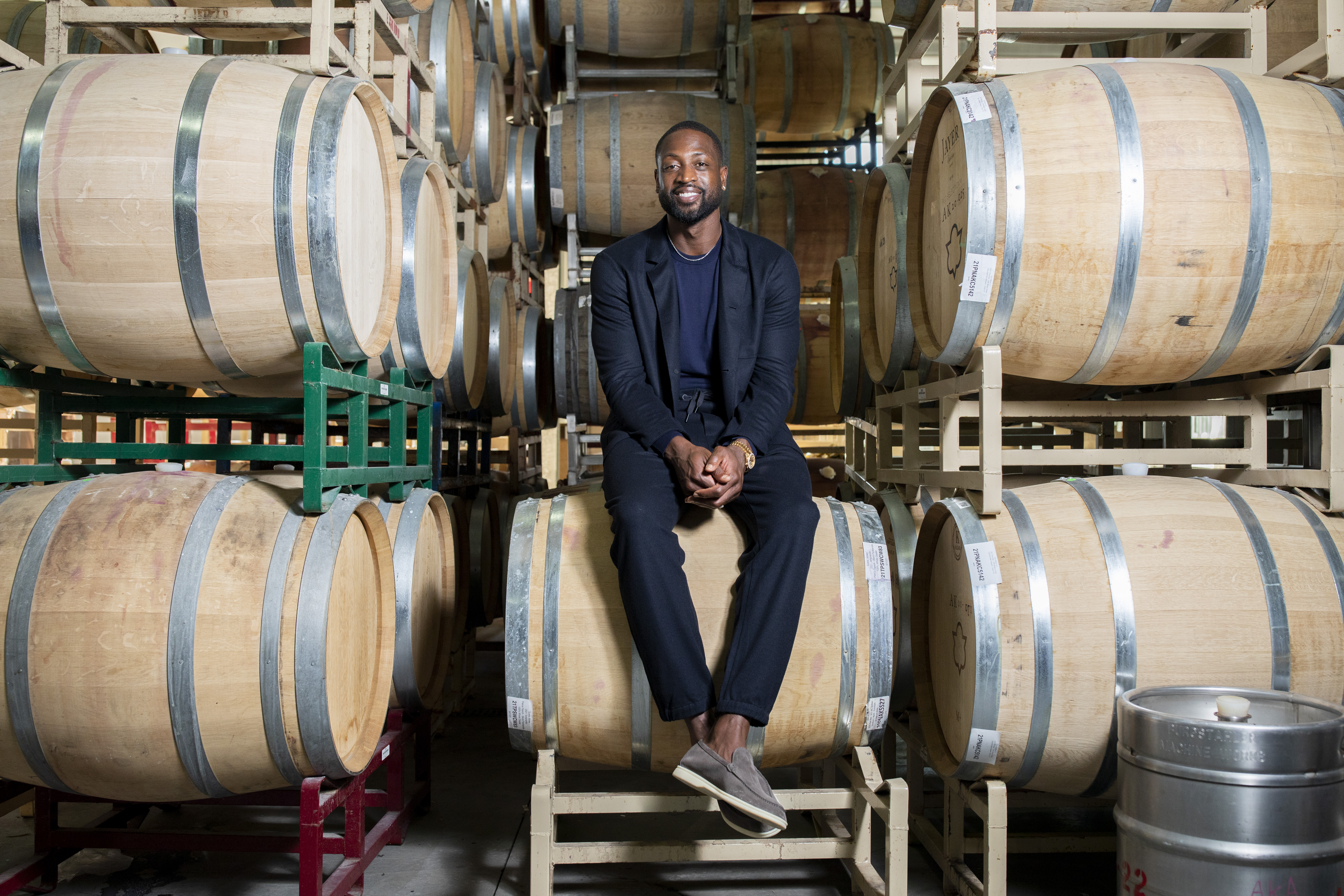 Tall man in a suit sitting in between a pile of stacked wine barrels. 