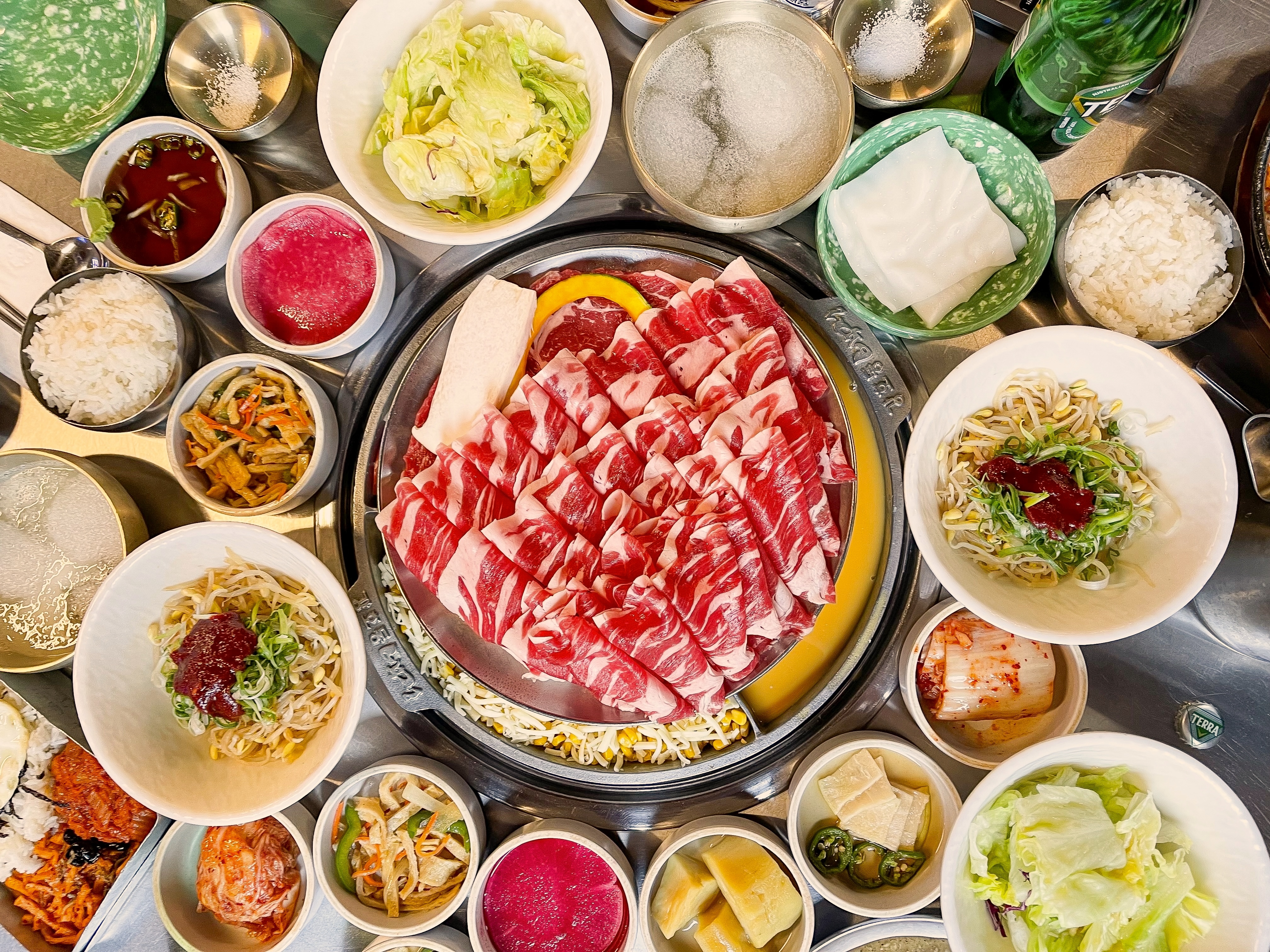 A pile of thinly sliced, highly-marbled beef surrounded by little bowls of noodles, raw vegetables, and various pickles.