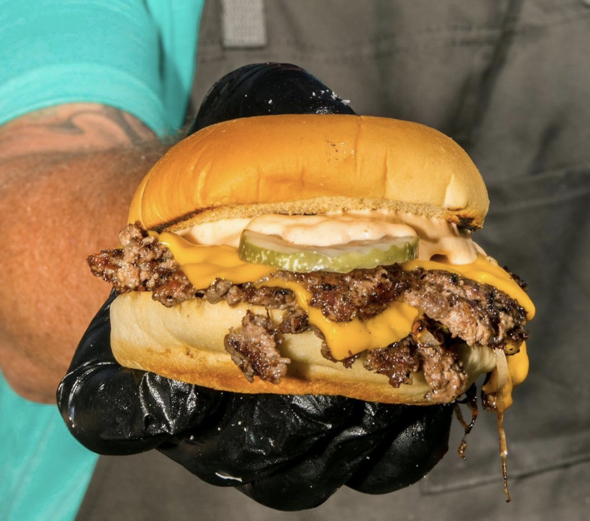 A hand in a black glove holds out a messy cheeseburger.