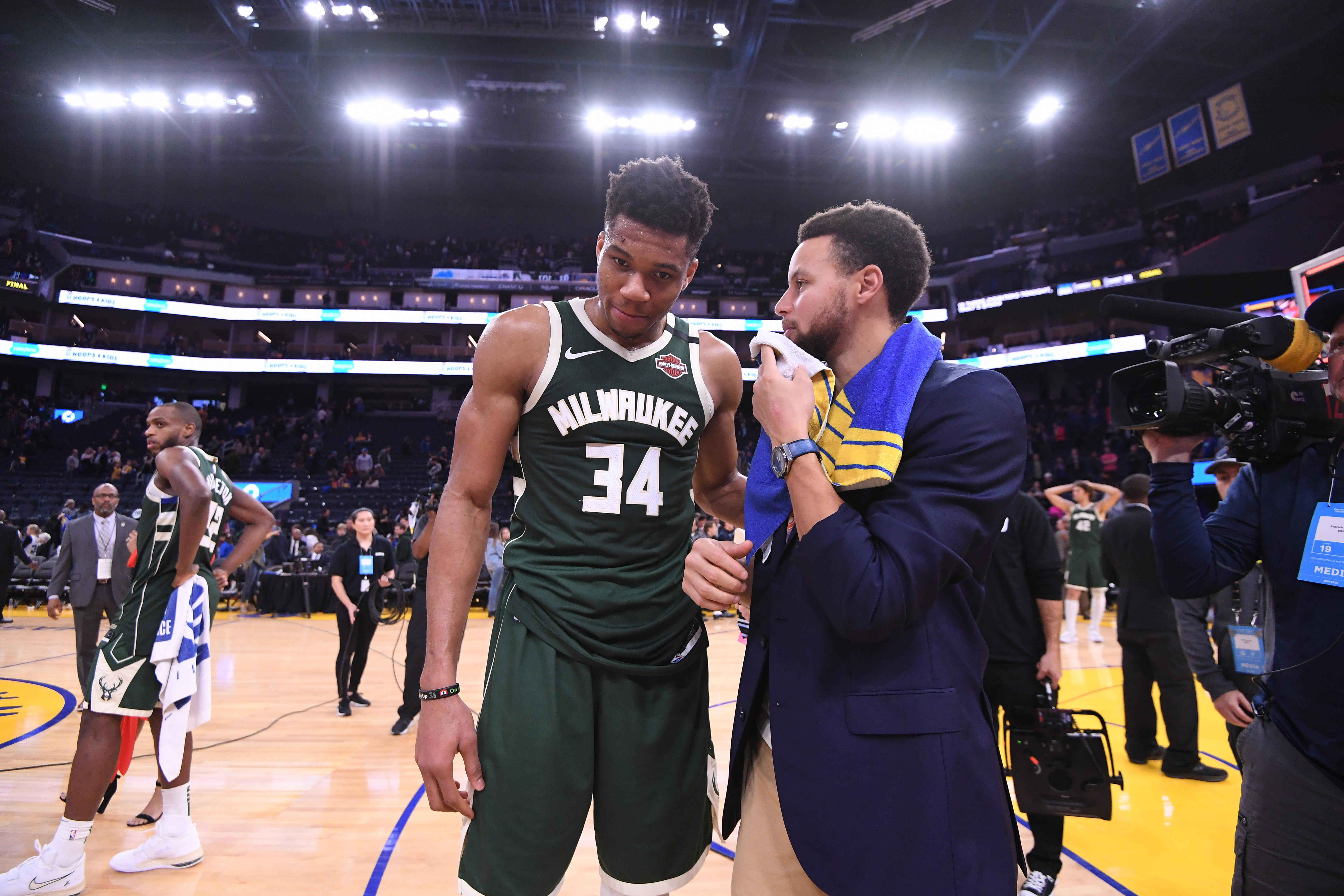 Steph Curry, in street clothes, covers his mouth while talking with Giannis Antetokounmpo after a game