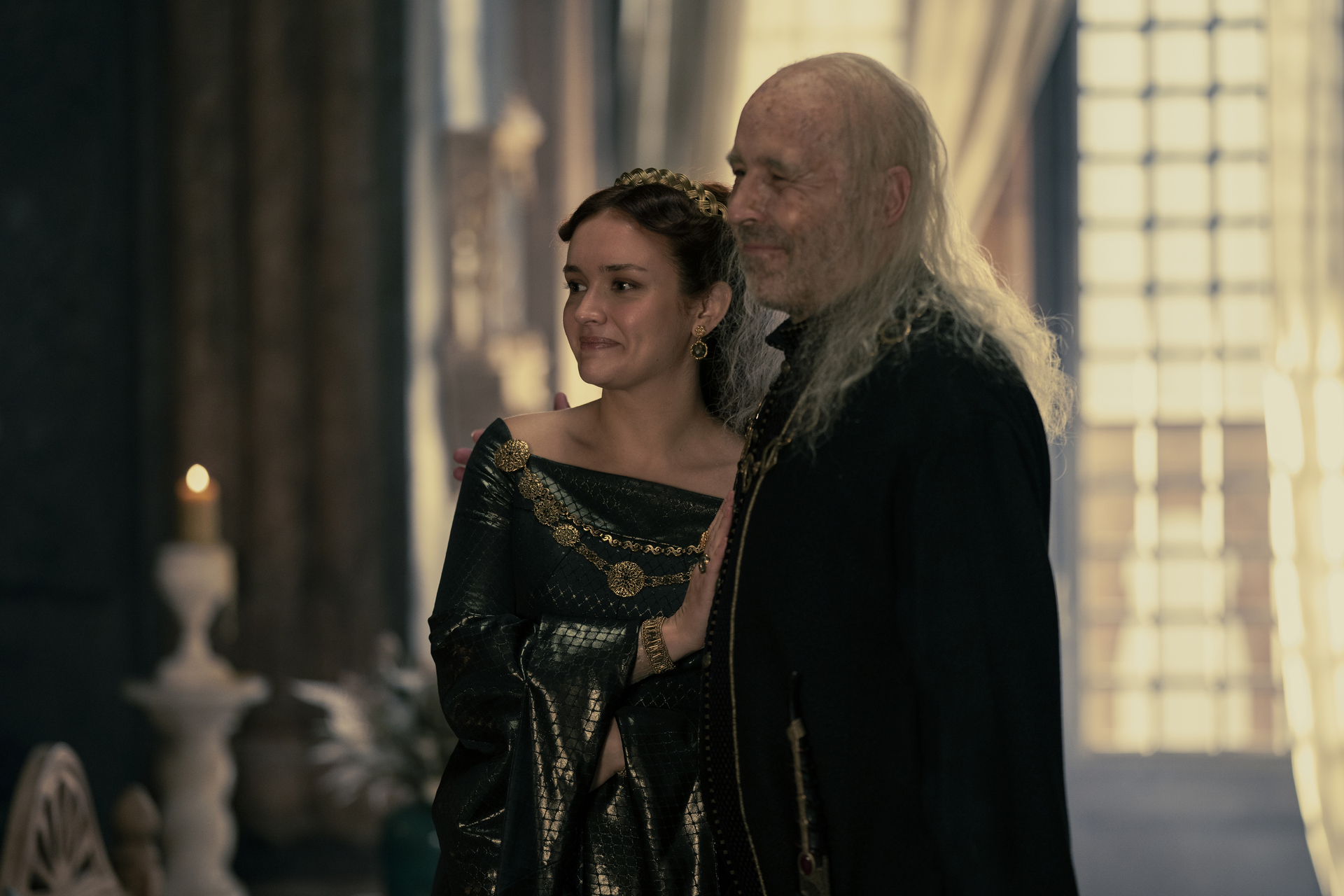 Alicent Hightower (played by Olivia Cooke) stands close to her husband Viserys Targaryen (played by Paddy Considine) resting her hand on his chest. Both are smiling, but Viserys looks quite sick. The image is from House of the Dragon episode 6.