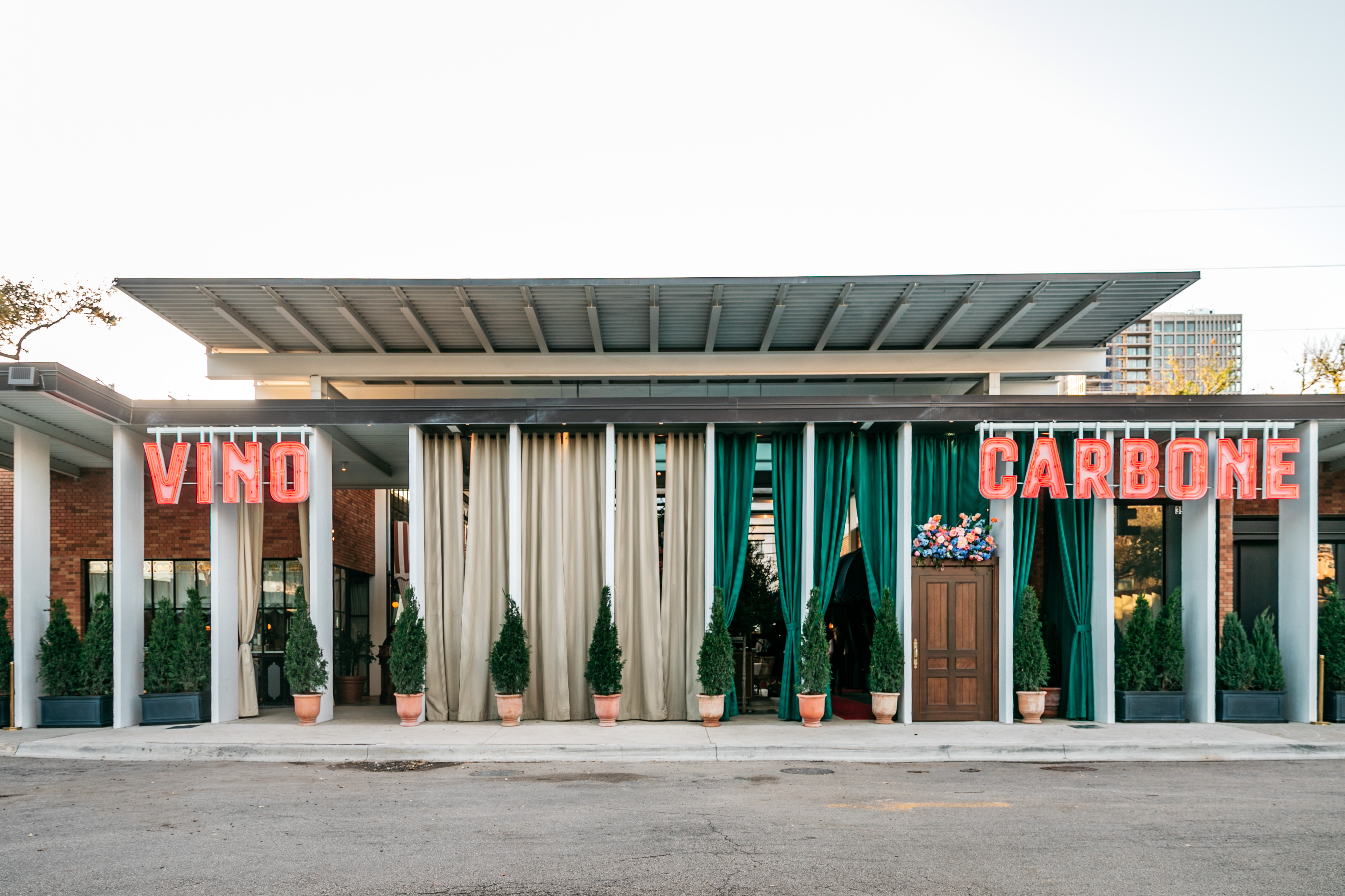 The exterior of Carbone Vino, a midcentury design building with all windows separated by partitions every few feet and an overhang. The neon sign reads in block letters Vino Carbone. A wooden door that’s slight off center marks the entrance.