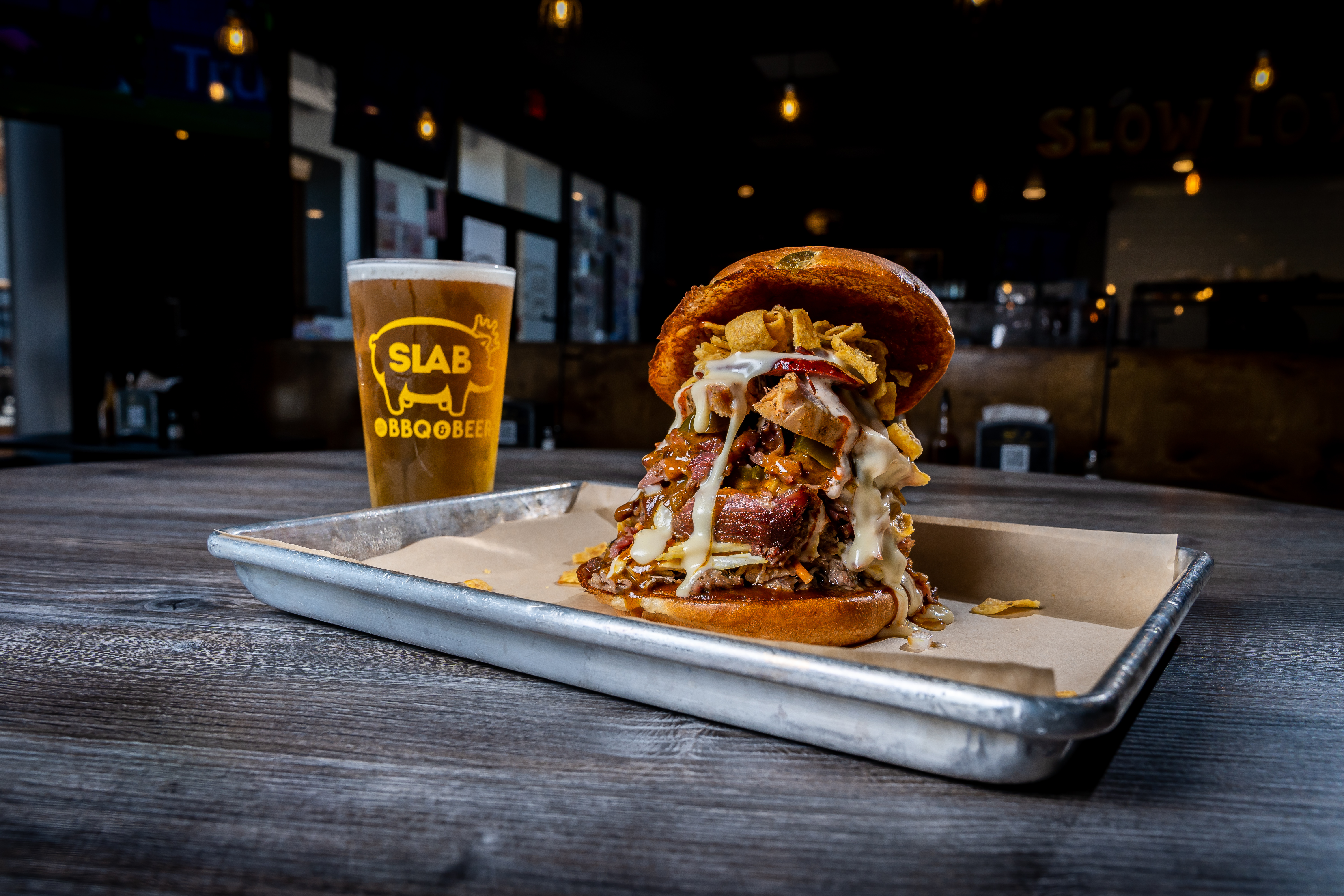 A tall sandwich with lots of meats and sauces and condiments on a tray next to a glass of beer with the words SLAB BBQ on it. 
