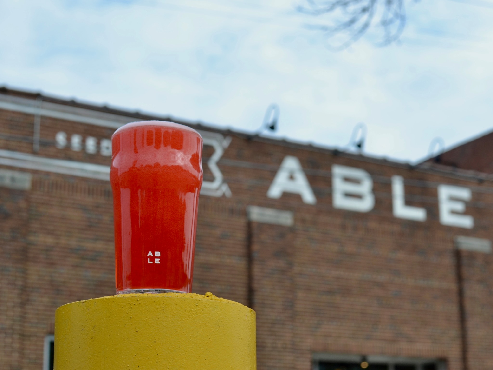 A reddish beer in a pint glass in front of a brick building with the word “Able” painted on it in white paint. 