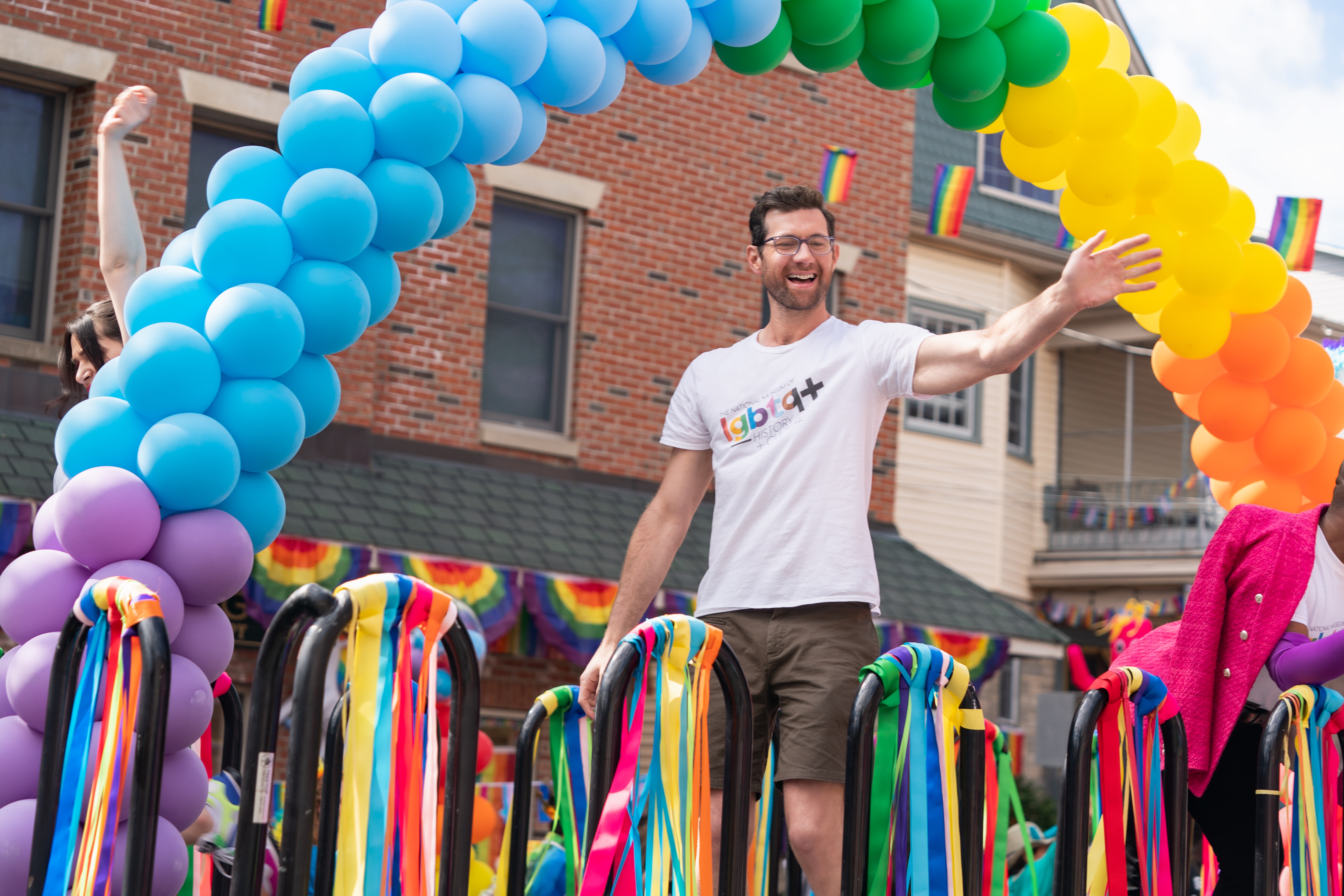 Bobby (Billy Eichner) stands on a parade float decorated with a rainbow balloon arch and rainbow streamers as he waves to the crowd in his “LGBTQ History” t-shirt