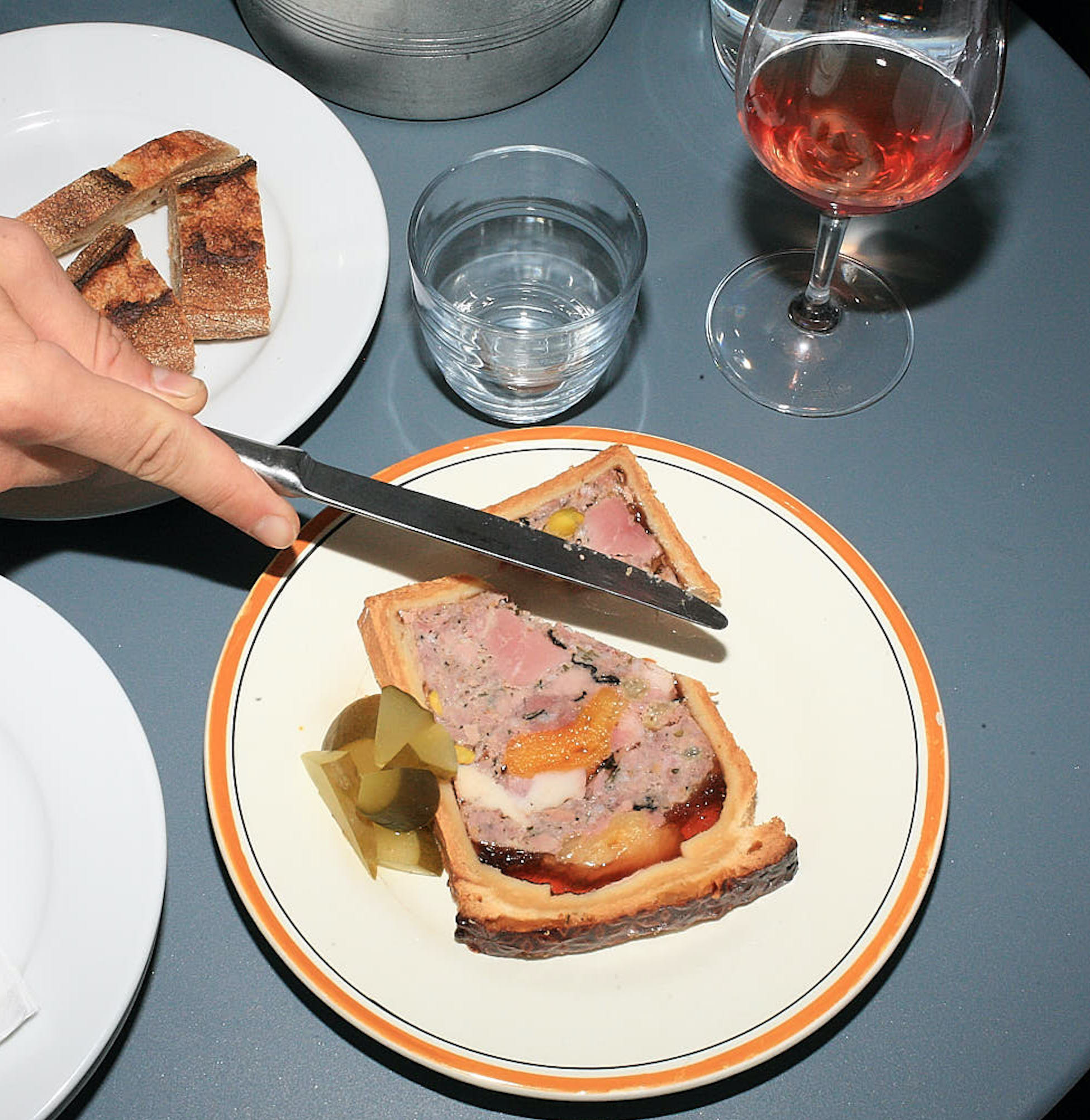 A hand cuts into a slice of patê en croute on a blue outdoor table, the kind seen at brasseries. A glass of pink wine and a plate of cut baguette sit alongside.