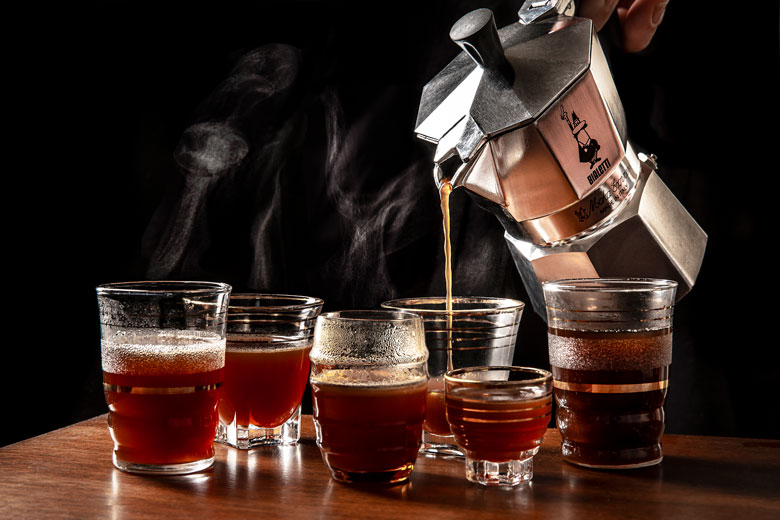 An array of clear glasses containing coffee and a coffee pot pouring coffee into one of them