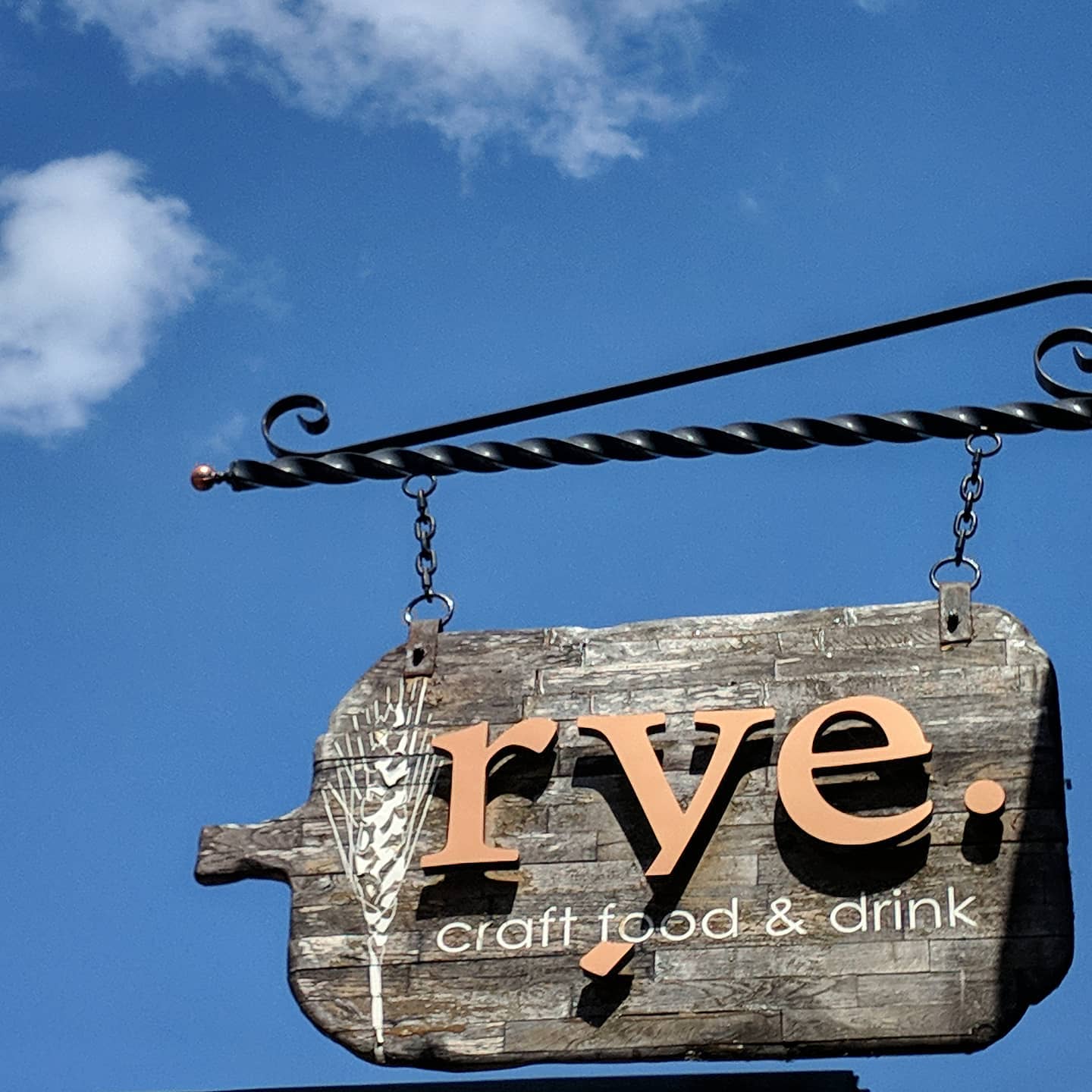 A wooden sign shaped like a jug hangs from chains. It reads: “Rye. Craft food and drink” in faded orange and white letters.