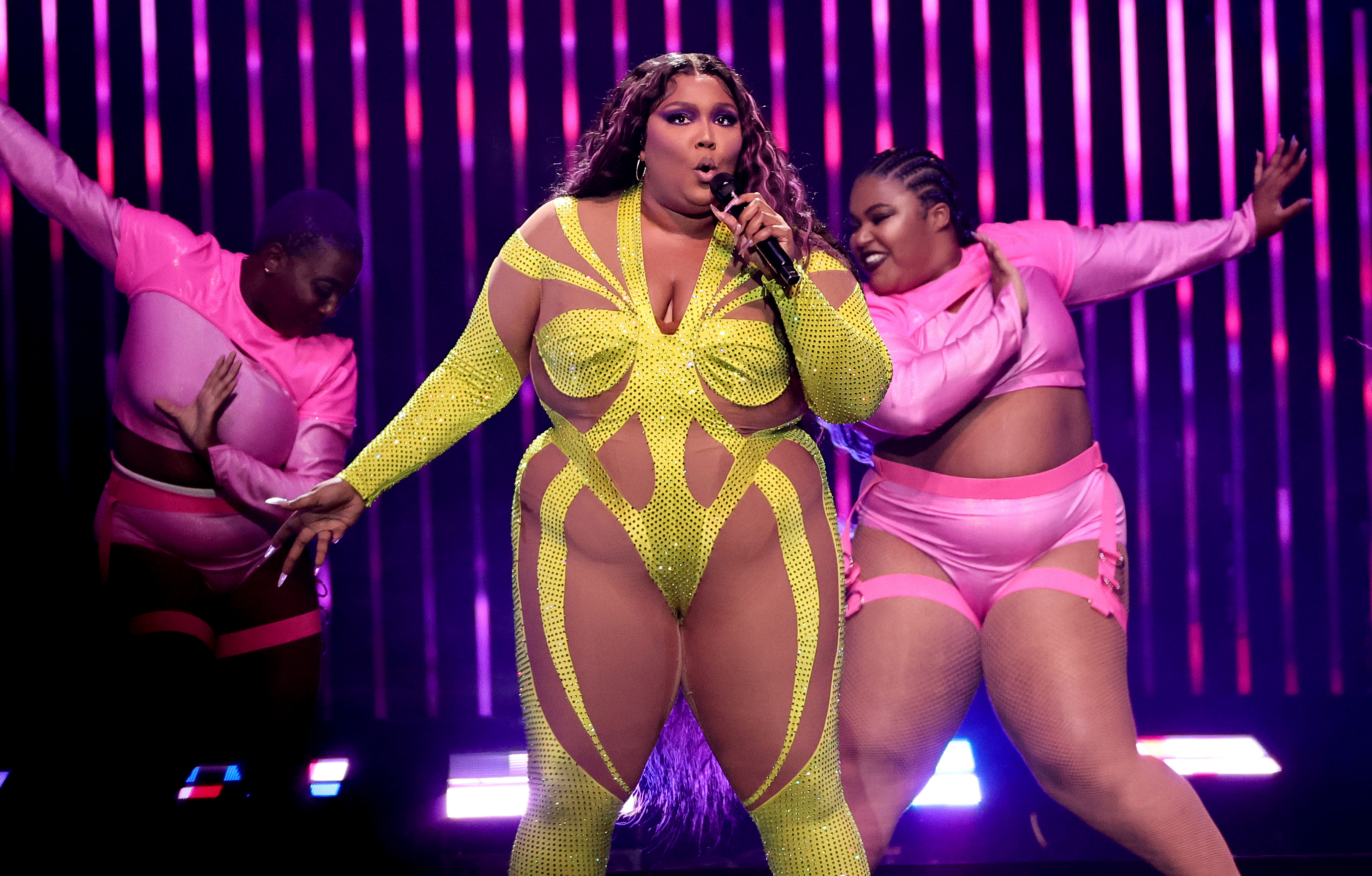 Lizzo singing live in a concert  in New York City. She is wearing a sheer and neon yellow body suit and is actively singing into the mike. She has dancers and pink lights behind her. 