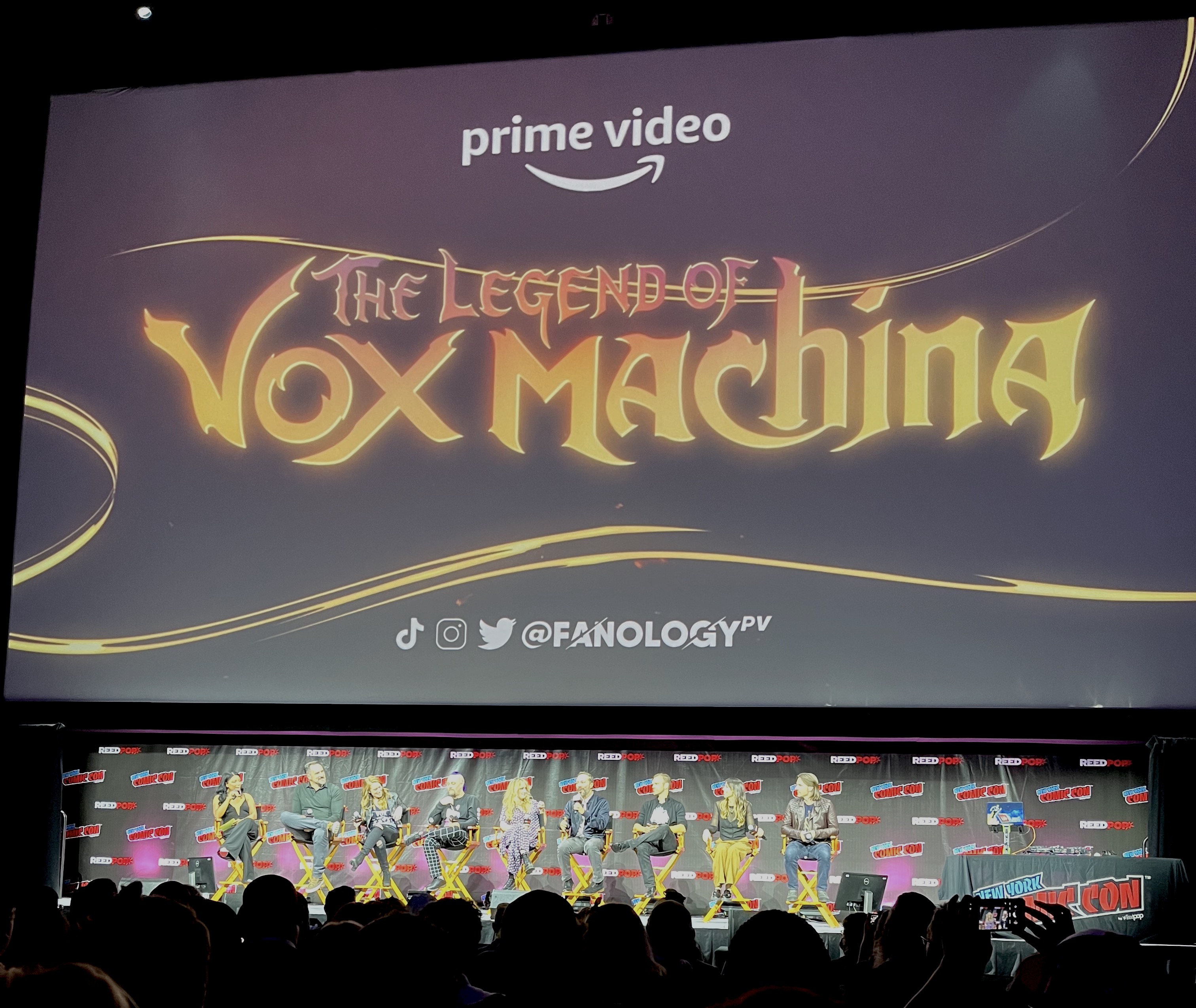 A picture of the New York Comic COn stage with the cast of Critical Role sitting in front of a giant screen projected with the “The Legend of Vox Machina” logo