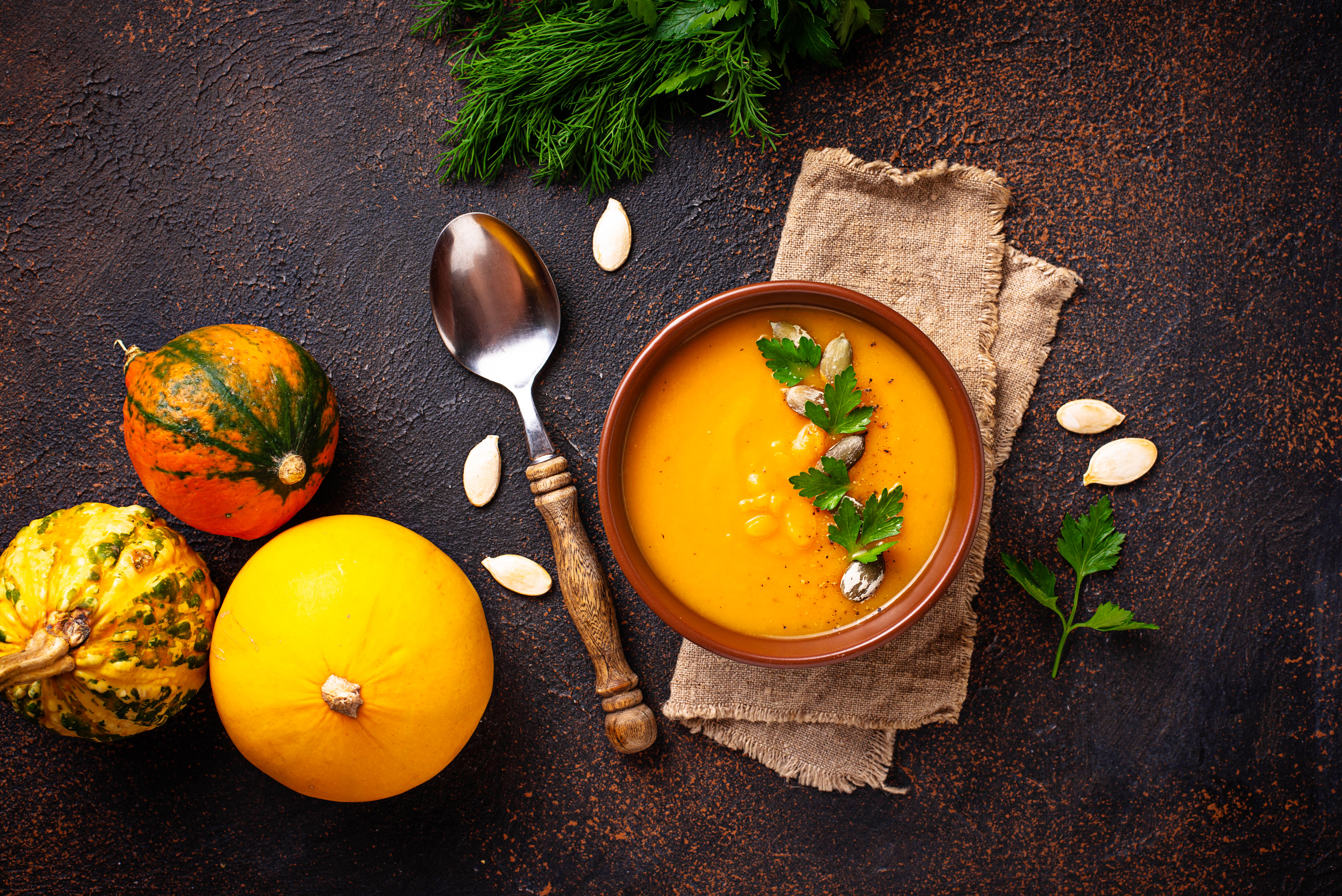 An overhead photo of a bowl of soup on a tan napkin with a spoon and three small pumpkins to its left.