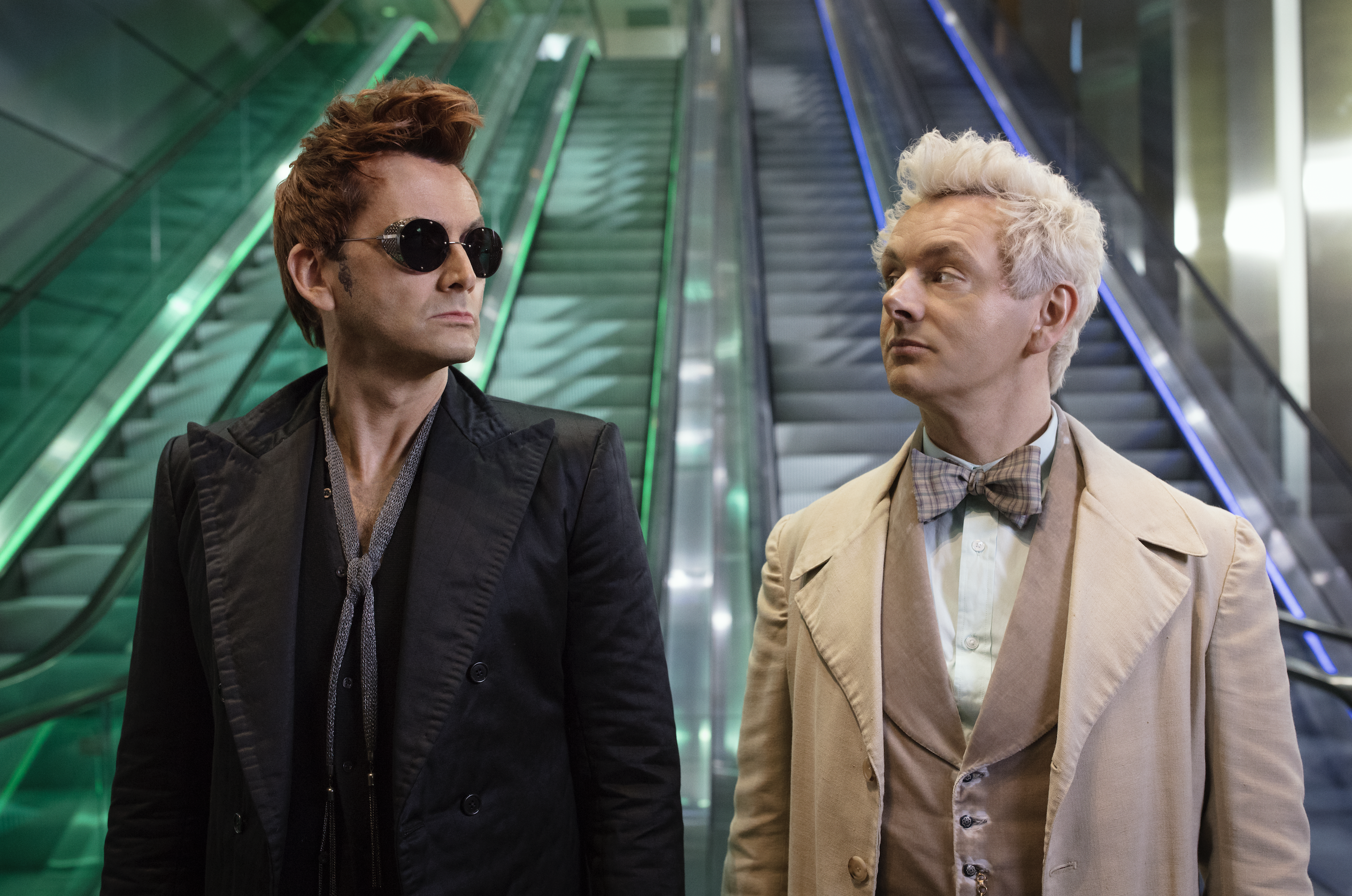 David Tennant as Crowley and Michael Sheen as Aziraphale looking at each other while standing in front of a bank of escalators in Good Omens