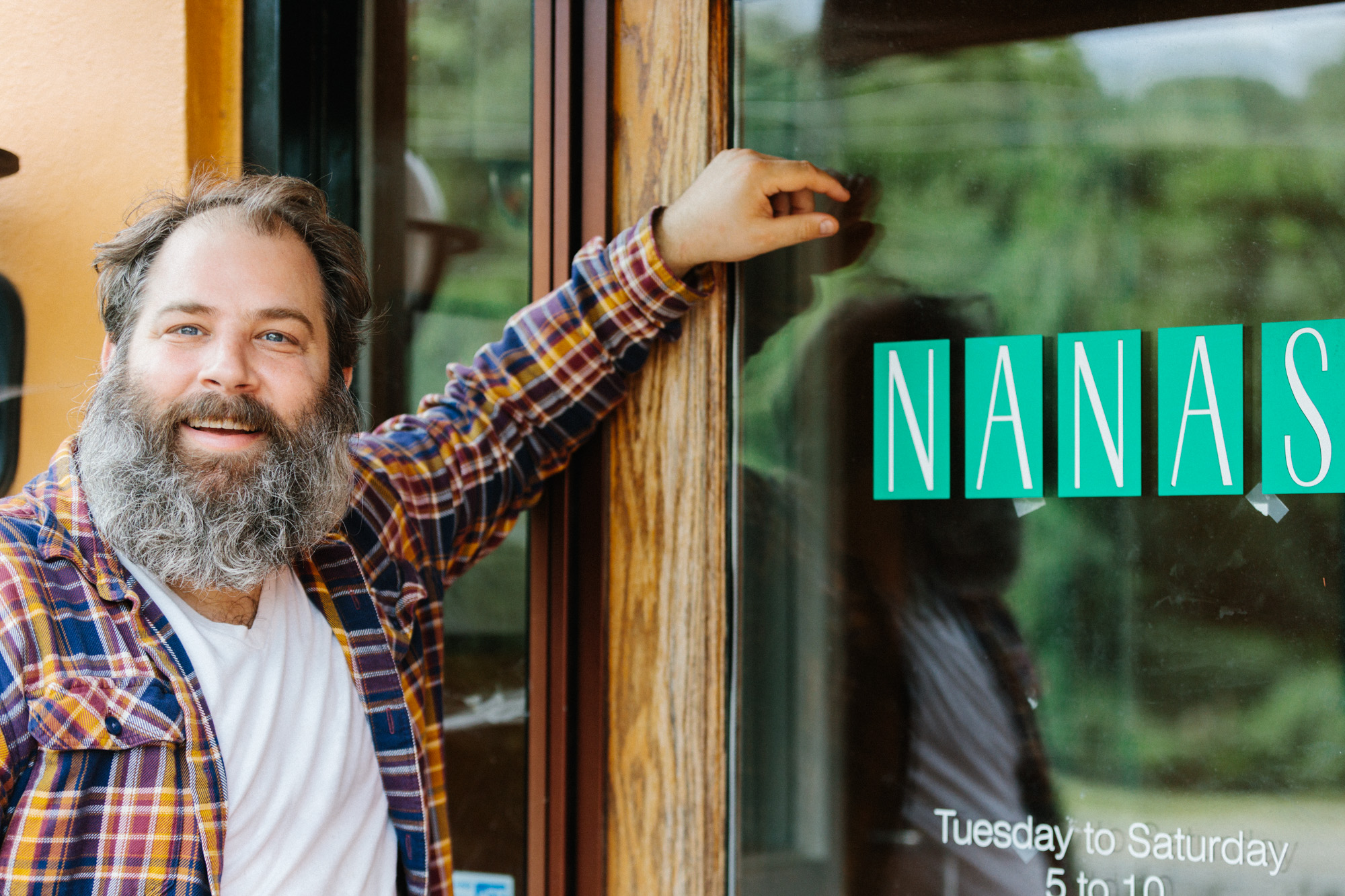 A white man with a beard leaning against a glass door that reads “Nana’s.”