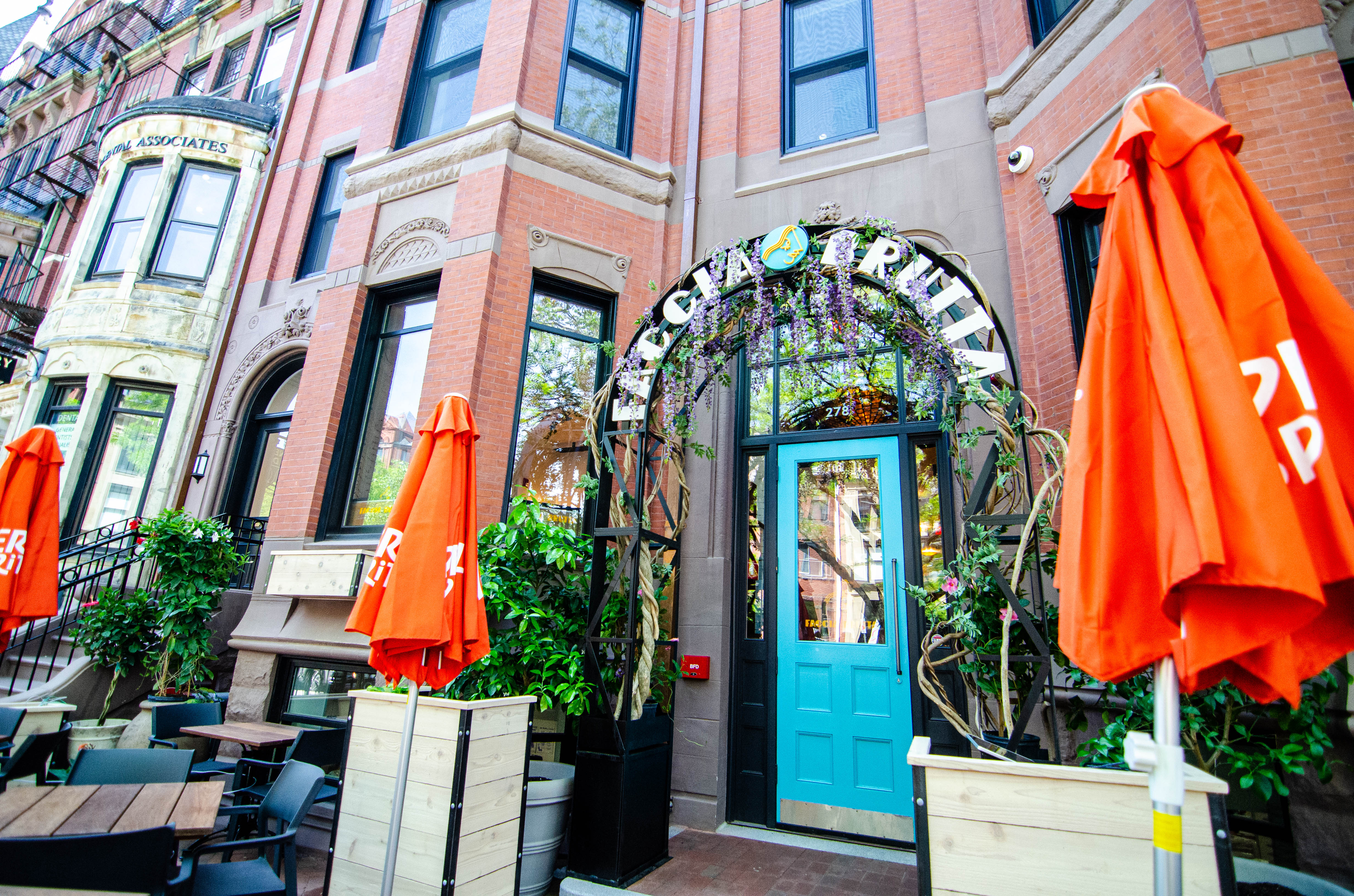 A restaurant exterior in a brick townhouse. A patio out front features wooden tables and chairs, bright orange umbrellas, and a trellis with the restaurant name, Faccia Brutta.