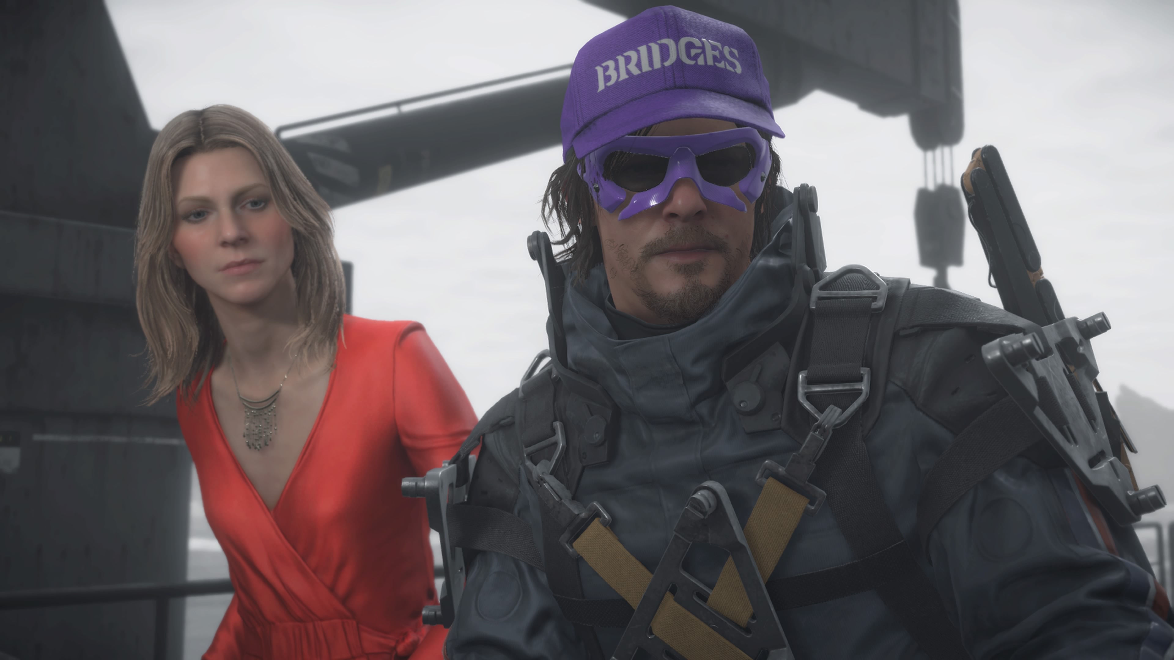 a middle-aged blond woman in a red dress standing next to a man in a futuristic spacesuit wearing a purple hat that says “BRIDGES” and purple-rimmed sunglasses, in Death Stranding