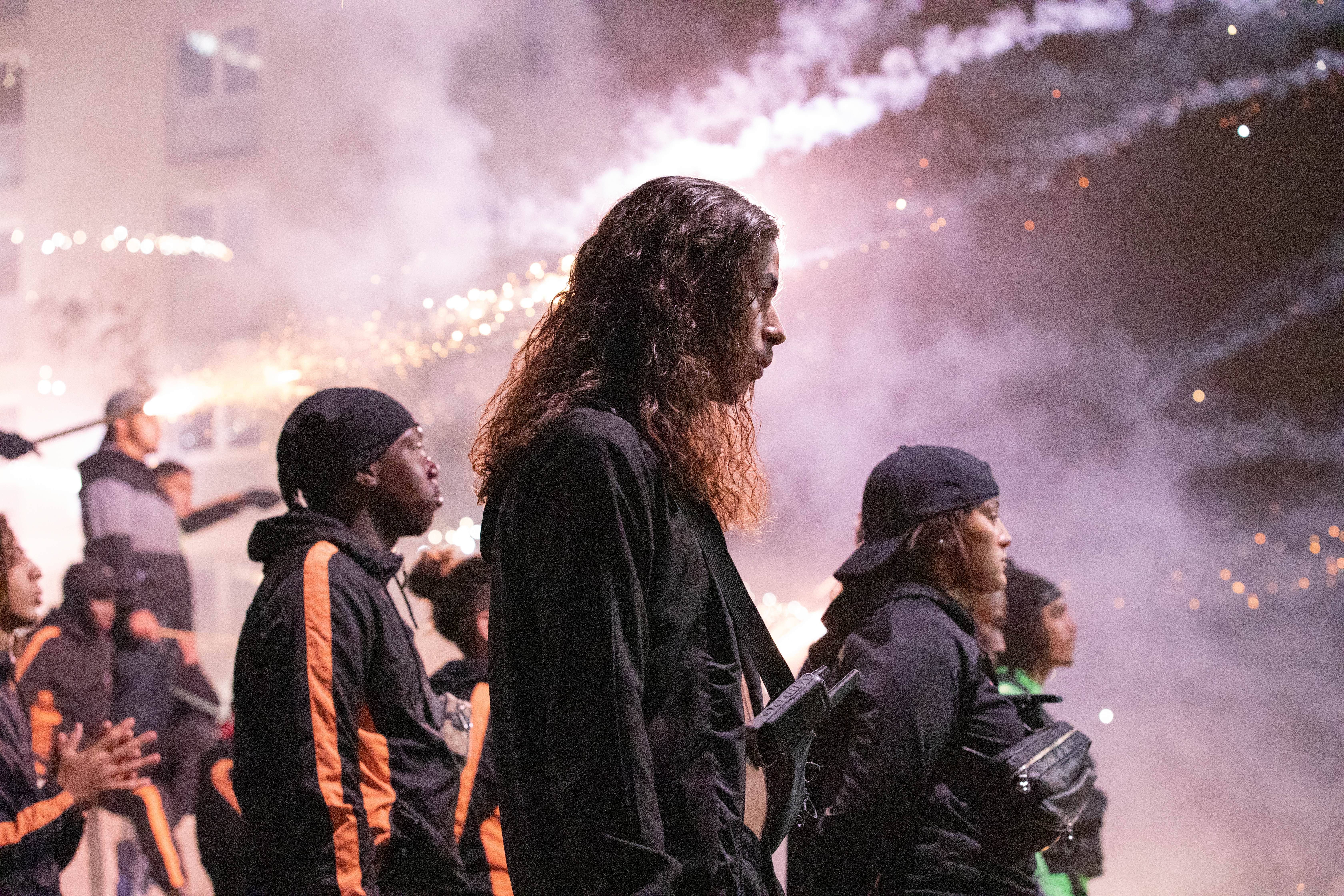 A side profile shot of a group of young men in black tracksuits with smoke and fireworks in the distance.