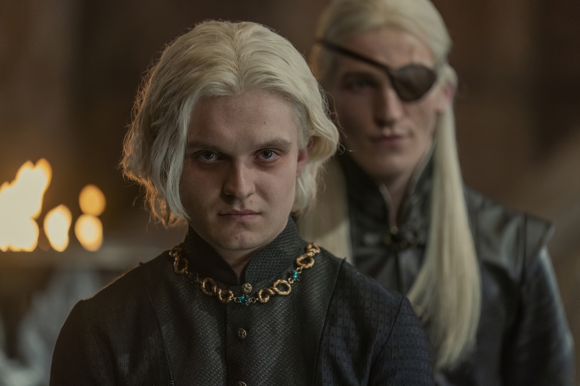 Aegon in the foreground looking grumpy about something, while his eyepatch-clad brother stands behind him out of focus, in House of the Dragon