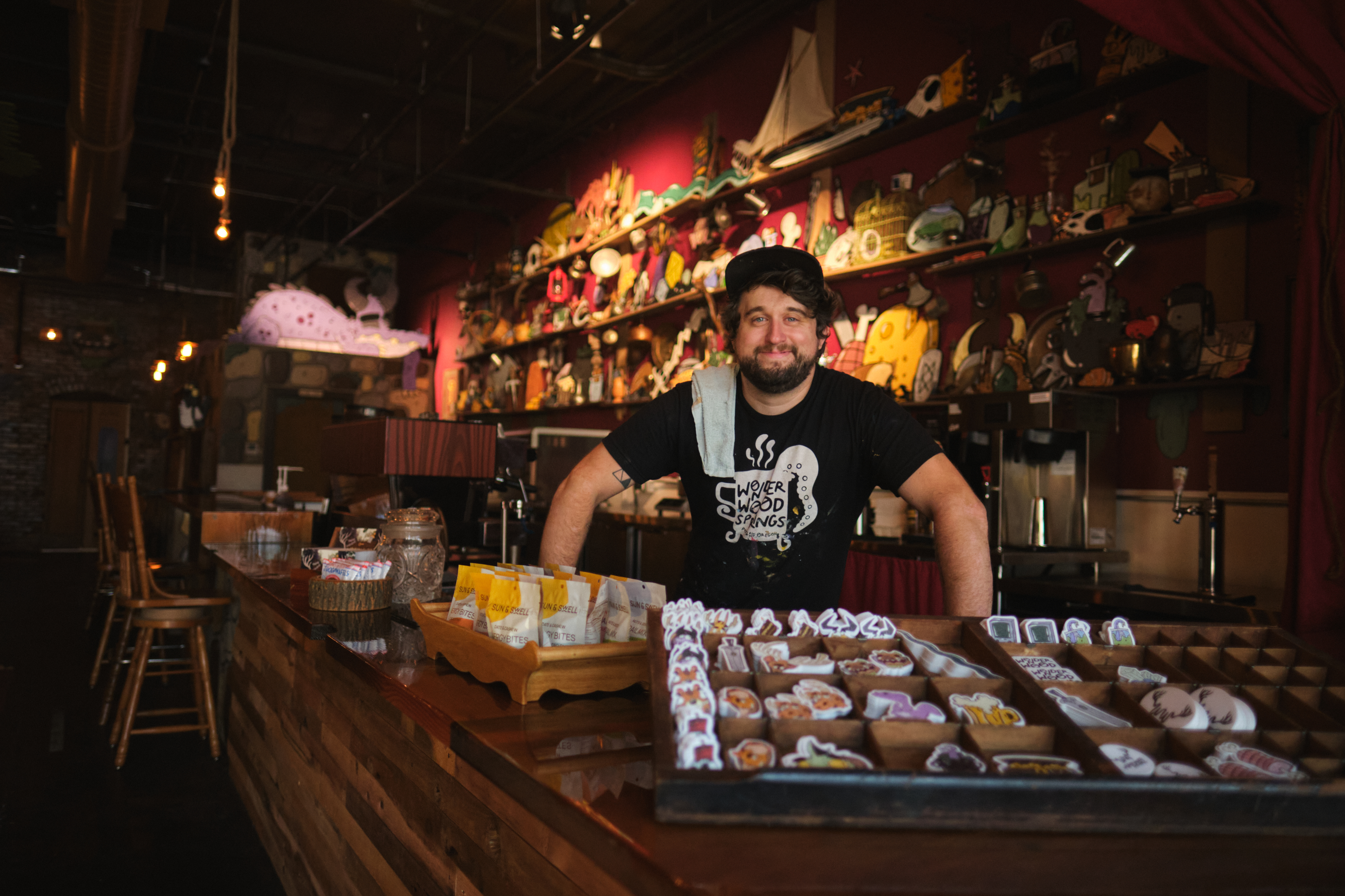 Mike Bennett stands behind the bar and in front of a large gallery wall of hand-painted art at his cafe.