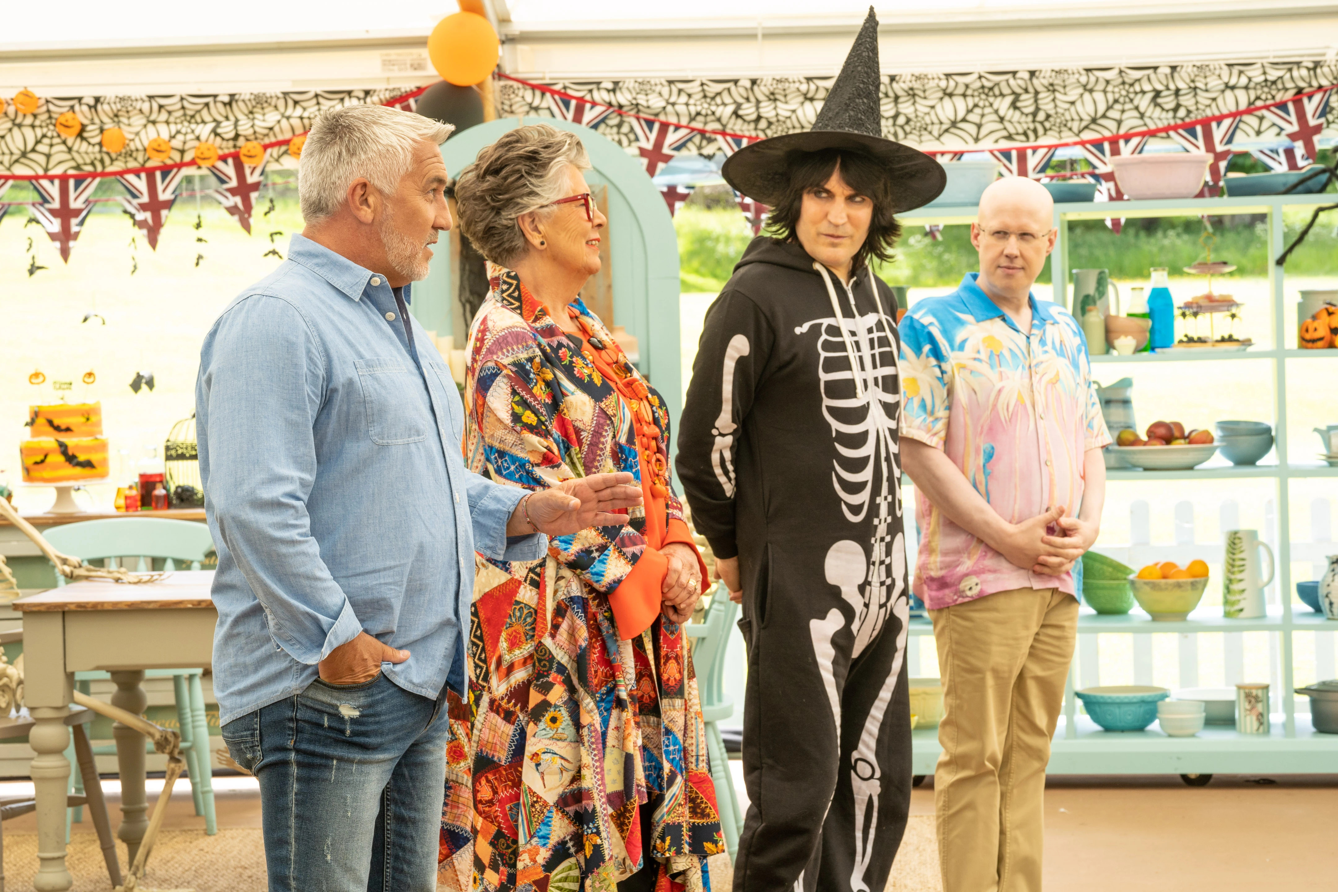 Four people stand in a tent adorned with pumpkins and cobwebs for Halloween Week; one, a man, wears a skull onesie and a witch’s hat.