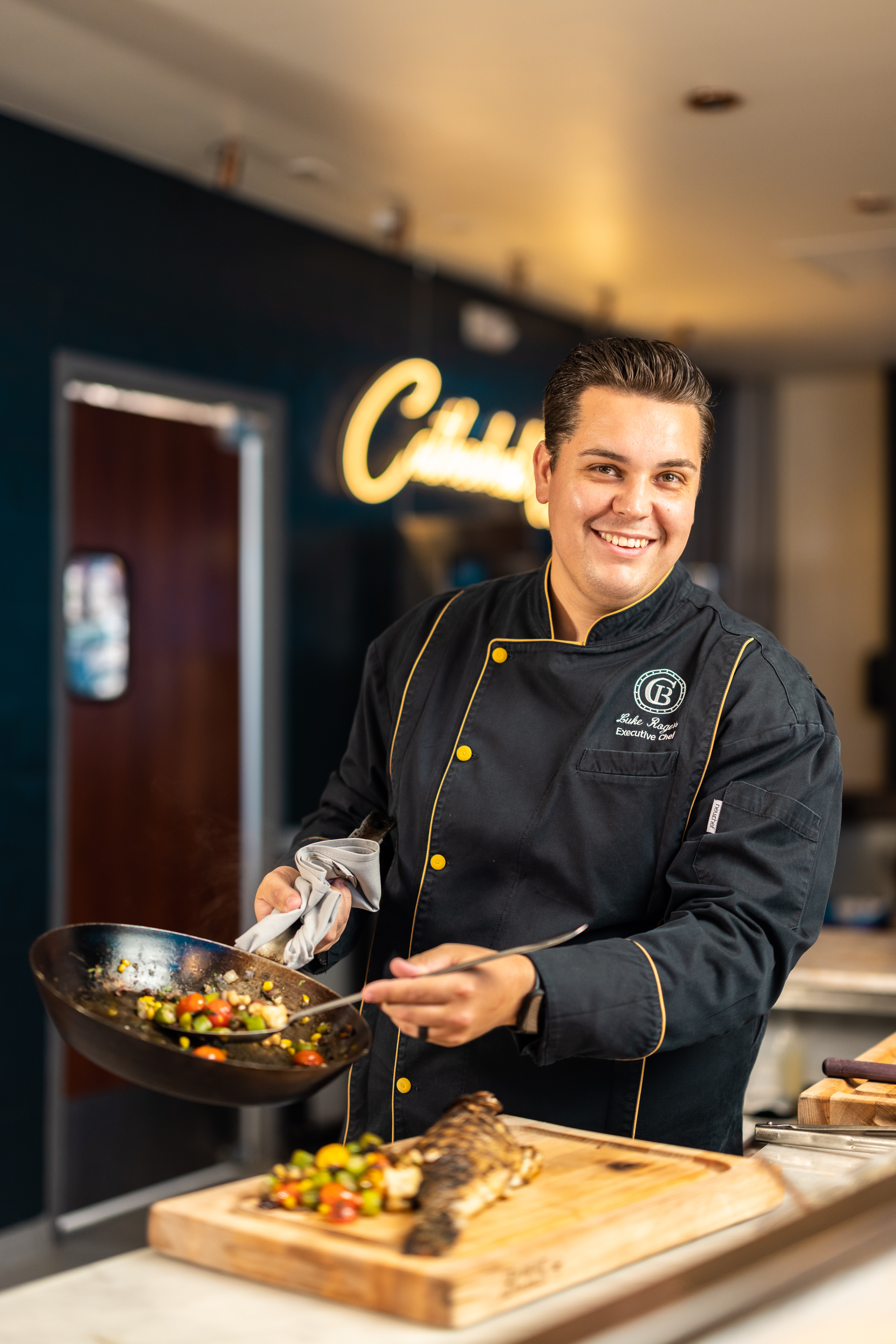 A chef in a black jacket holds a wok with cooking vegetables and smiles directly at the camera. 