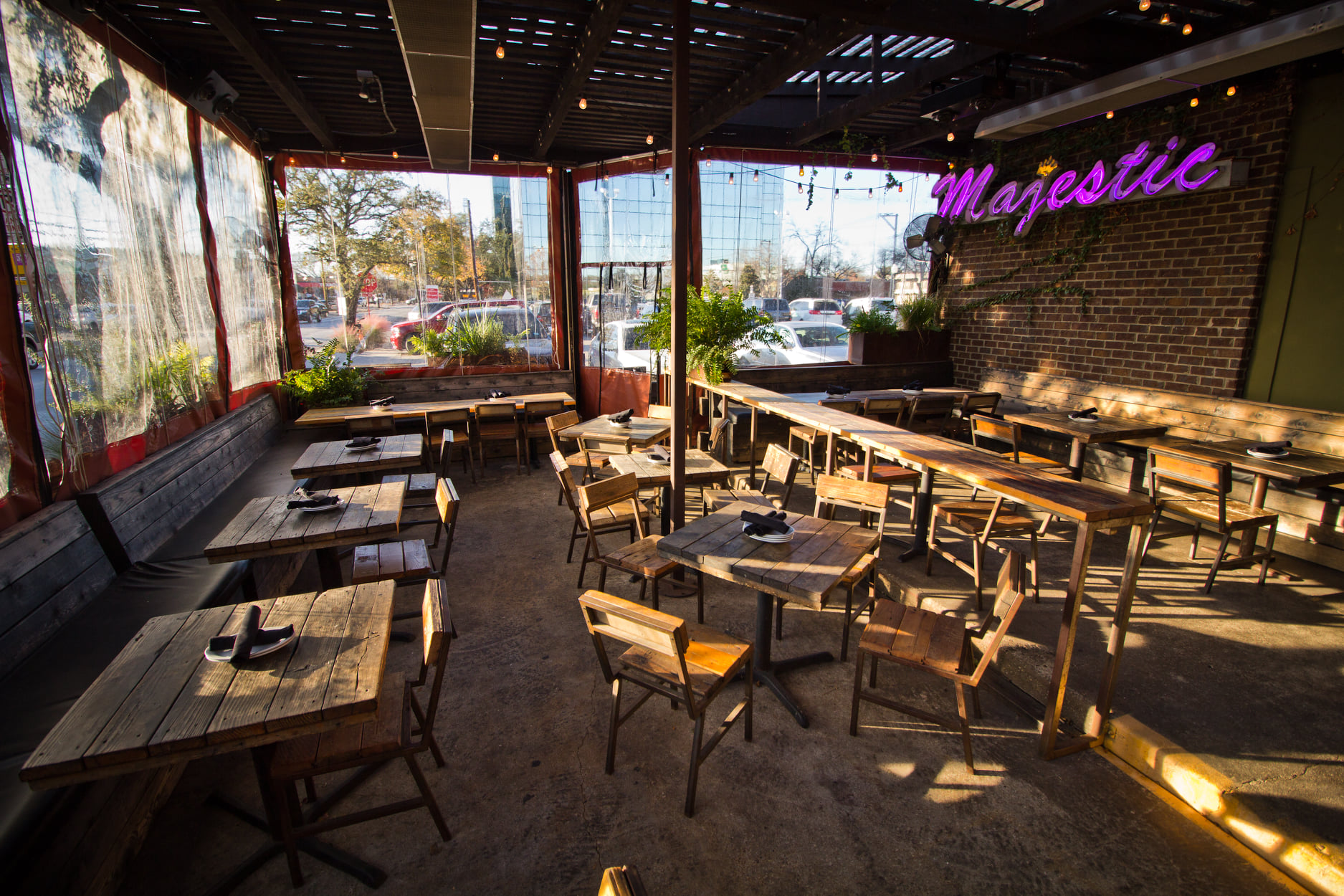 A shaded outdoor patio is furnished with wooden tables. In the background, a pink neon sign reads, “Majestic.”
