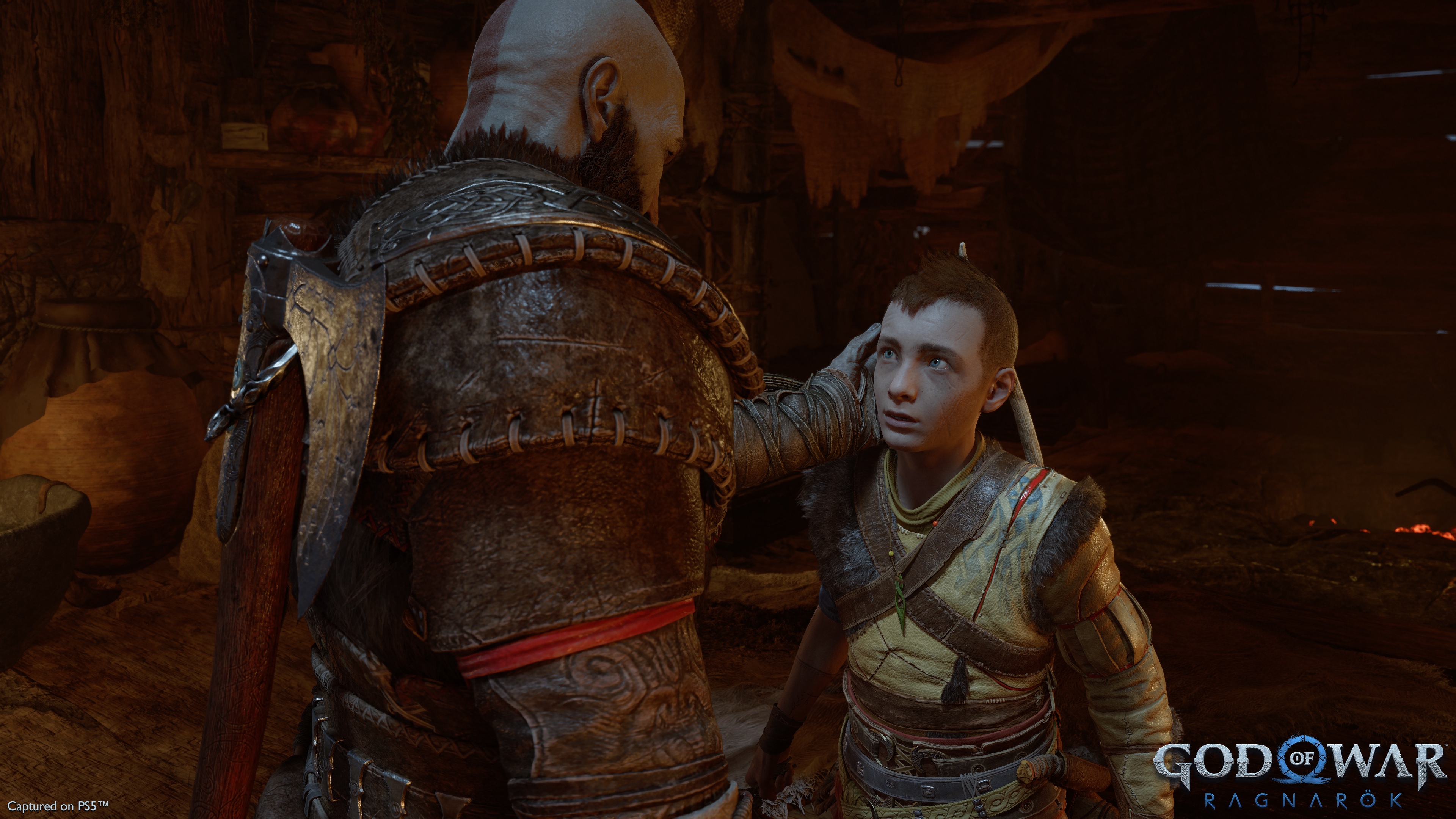 Kratos touches the side of Atreus’ head, who looks up at him, while the two are in their cabin, in a screenshot from God of War Ragnarok.