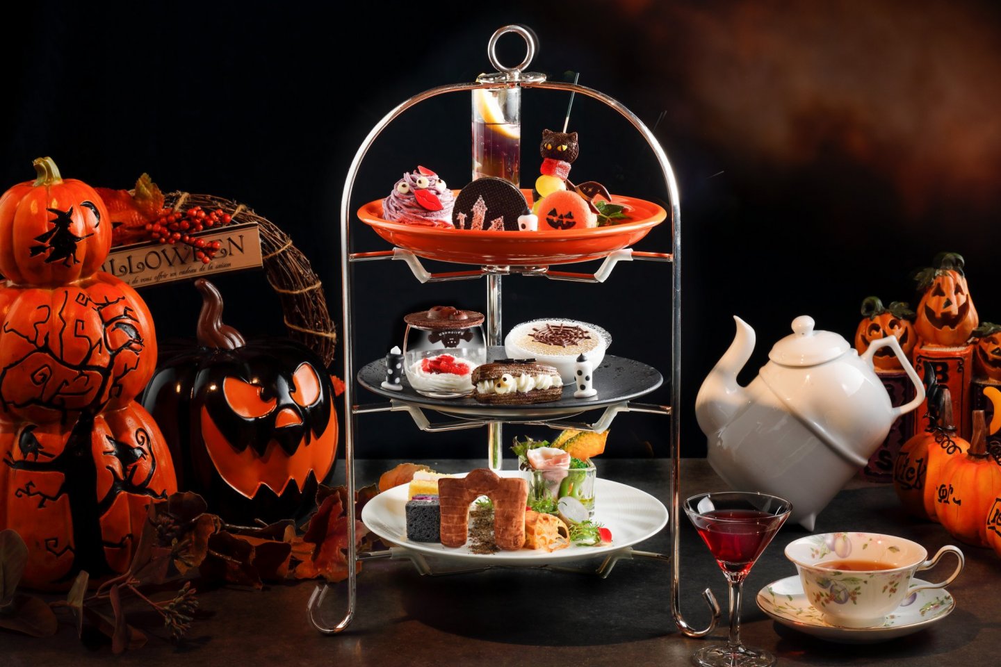 An afternoon tea set with three-tiered tray with Halloween cookies and candies, and finger sandwiches. White teapot set and pumpkins surround it.