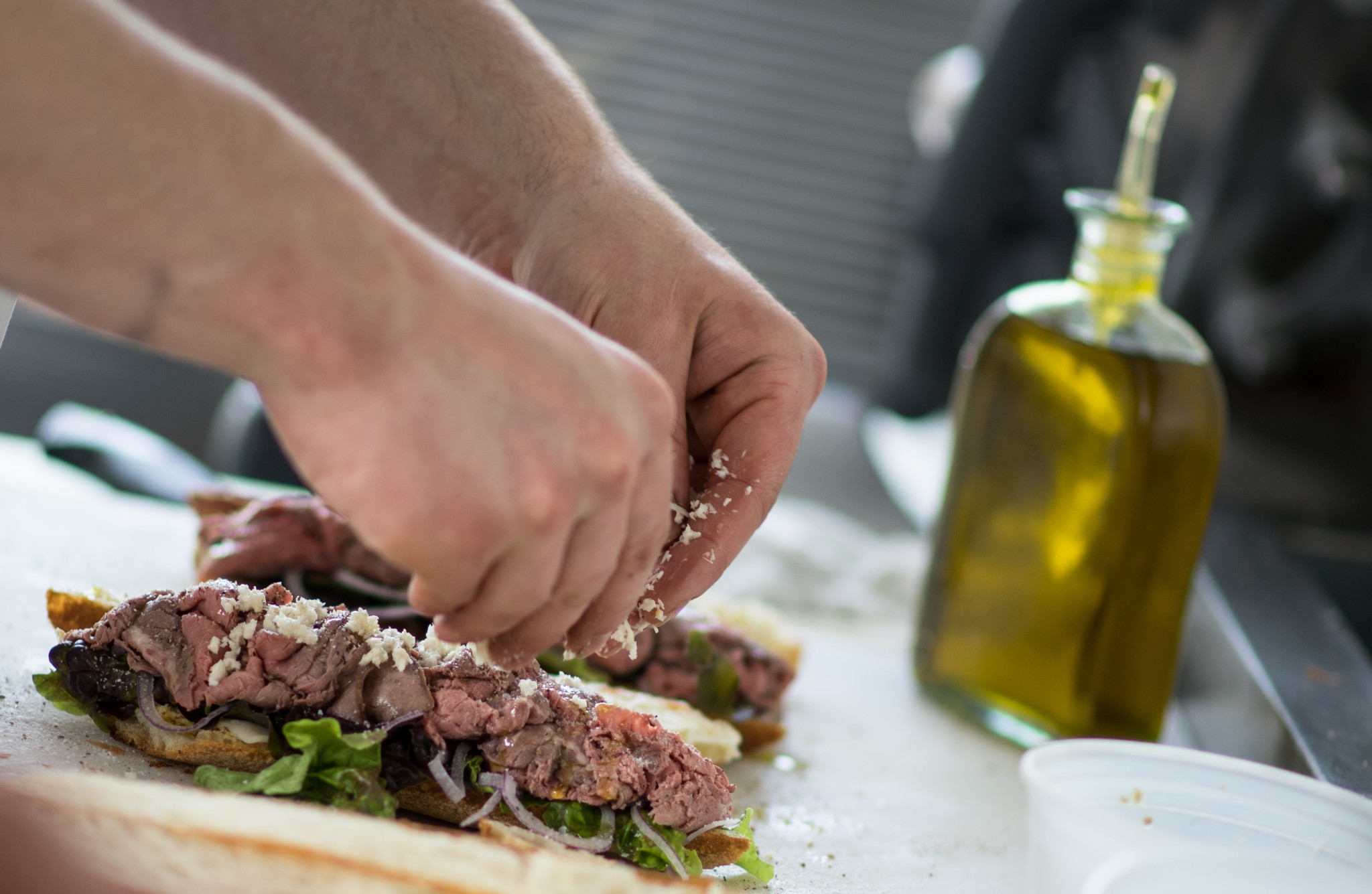 Two hands place shaved beef, lettuce and cheese on a baguette. A bottle of olive oil is visible in the background. 