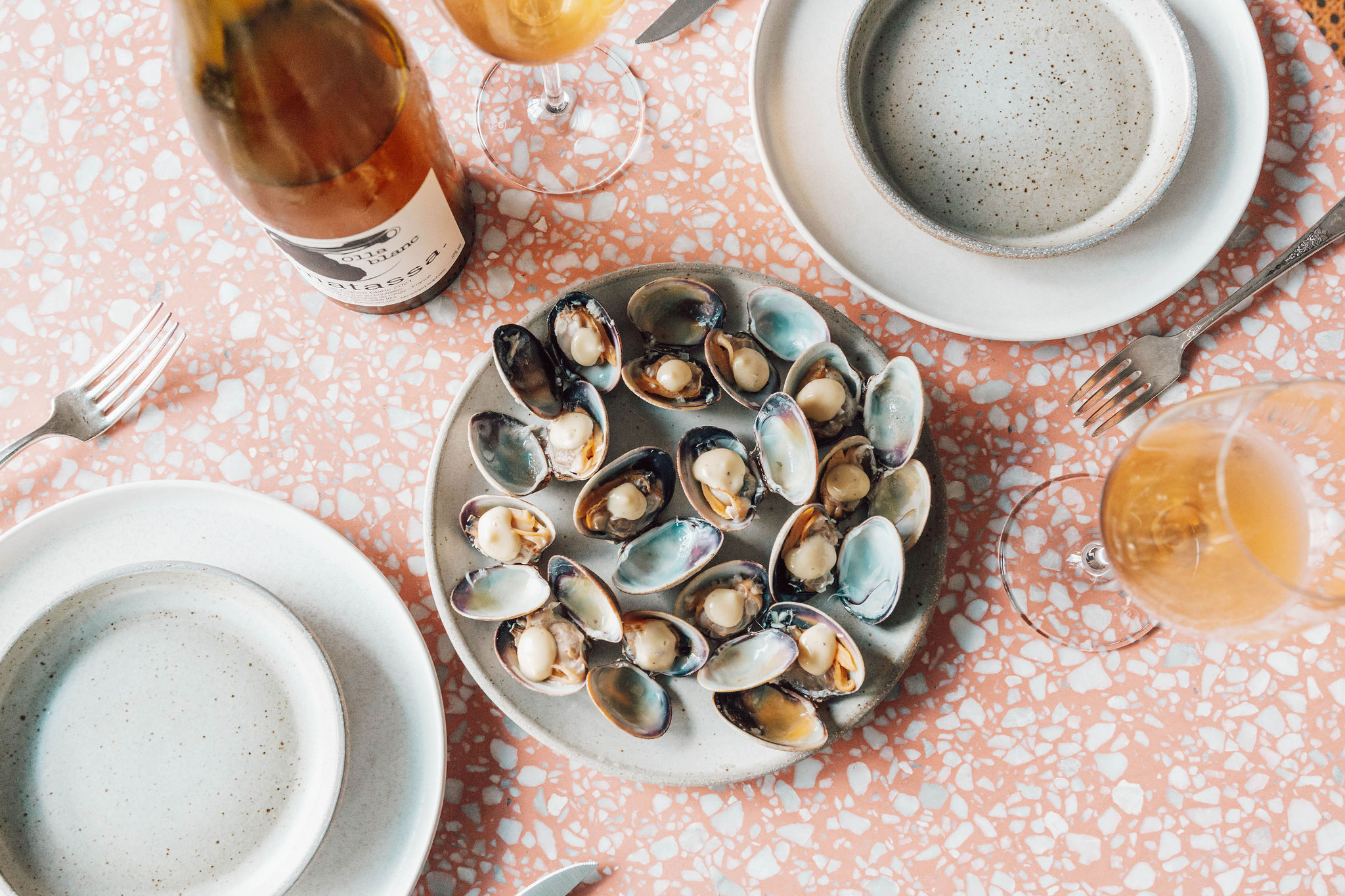A plate of open clams sits at the center of a table next to two glasses of orange wine at Heavenly Creatures.
