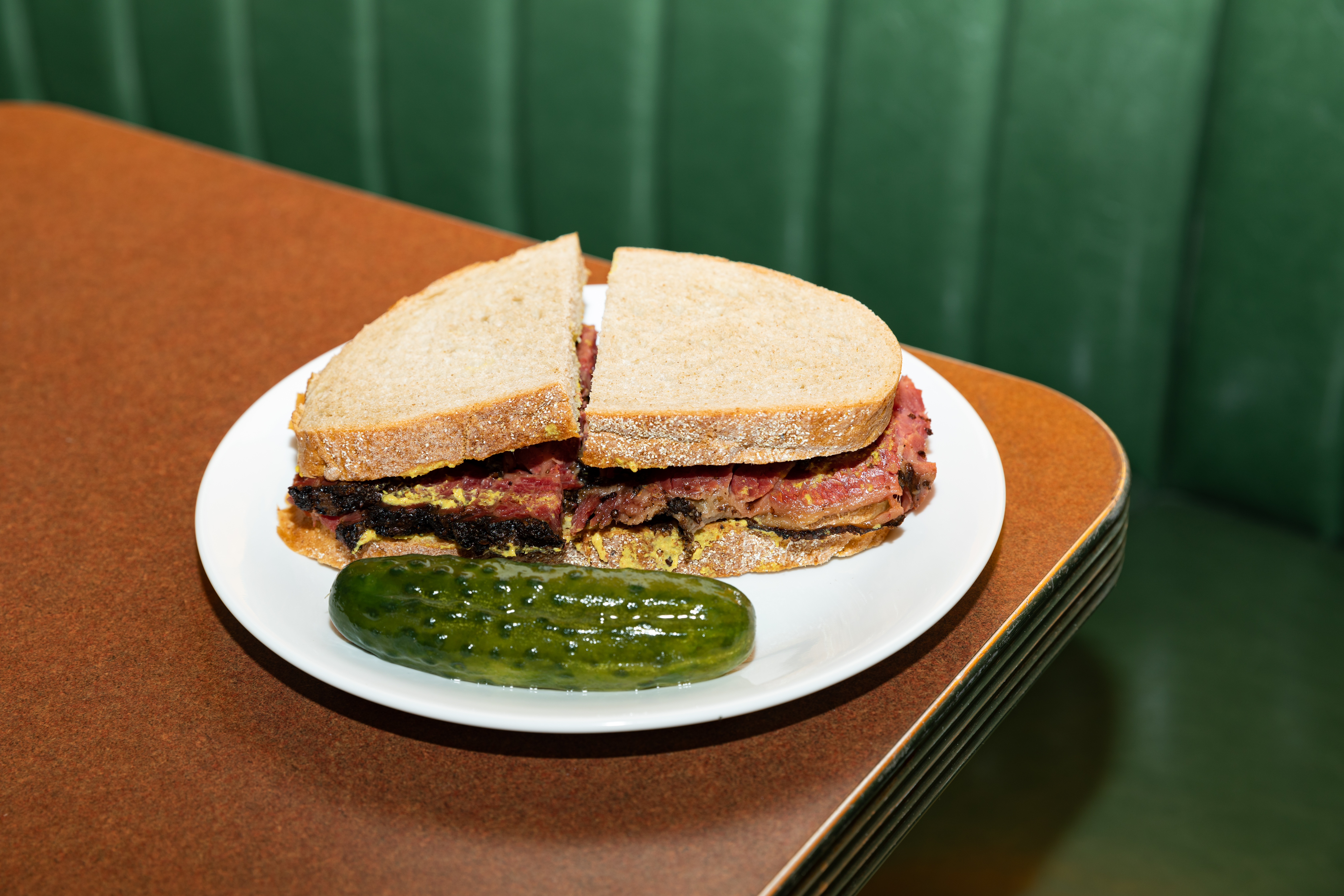 A mustardy pastrami sandwich on rye bread is served on a plate with a half-sour pickle.