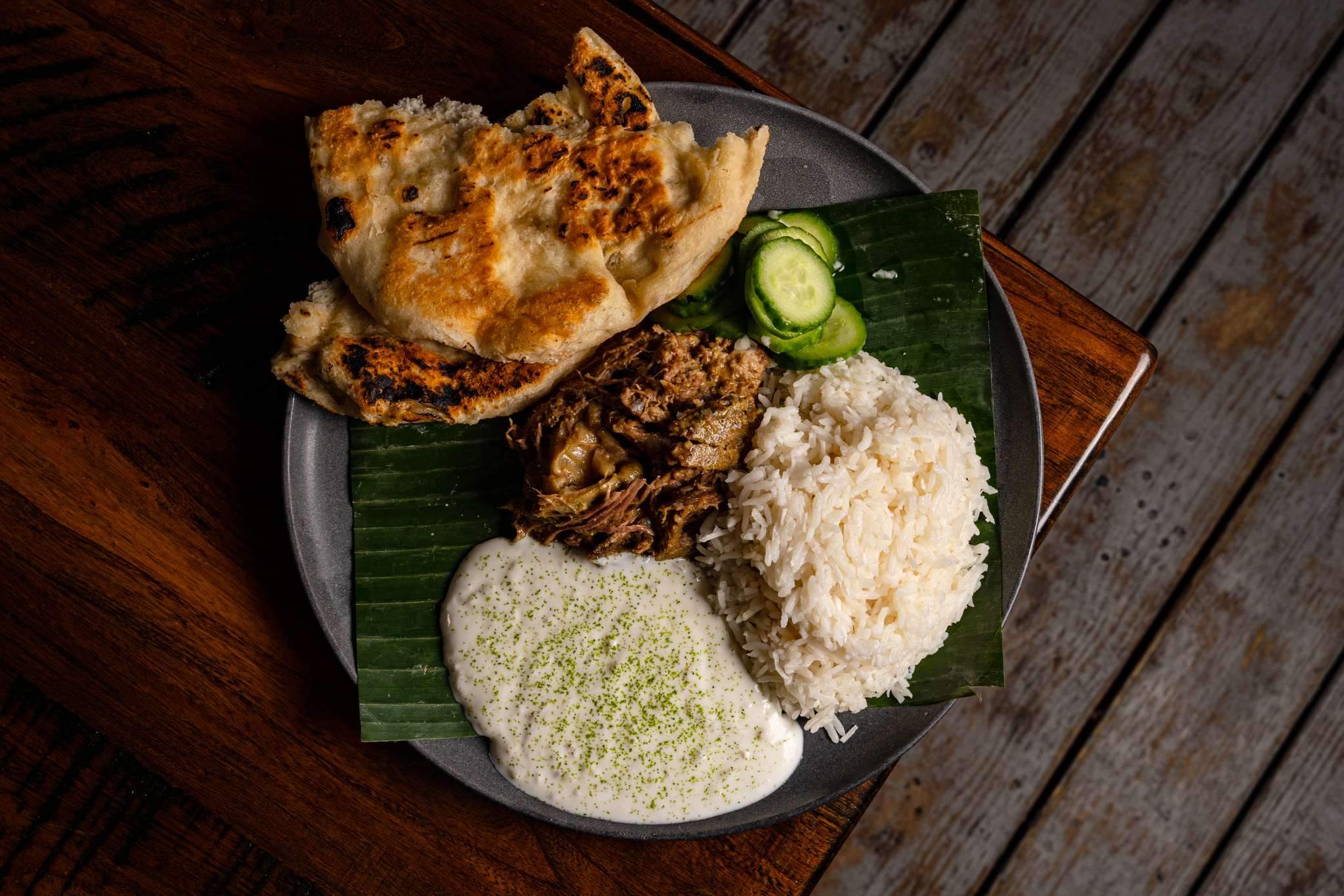A dish with a banana leaf, Indian bread, rice, meat, cucumbers, and a white sauce on a dark table