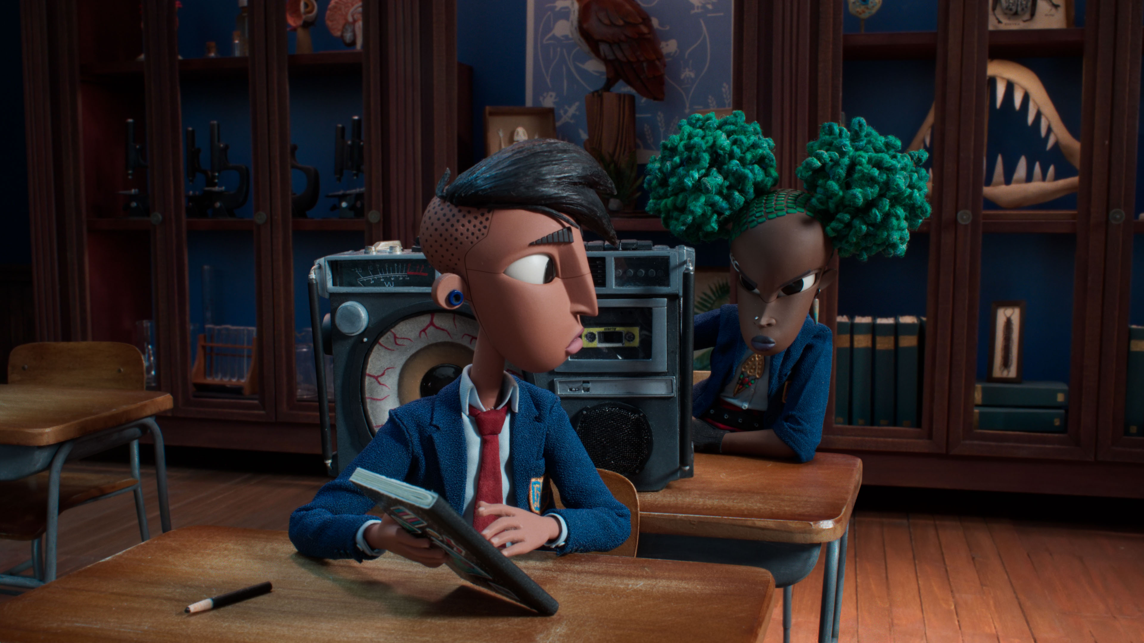 Raúl, a trans Latinx teenager with an undercut, sits in a school classroom at a desk looking back at Kat, a Black 13-year-old punk girl with bright green hair and a giant boombox, in the stop-motion animated film Wendell &amp; Wild