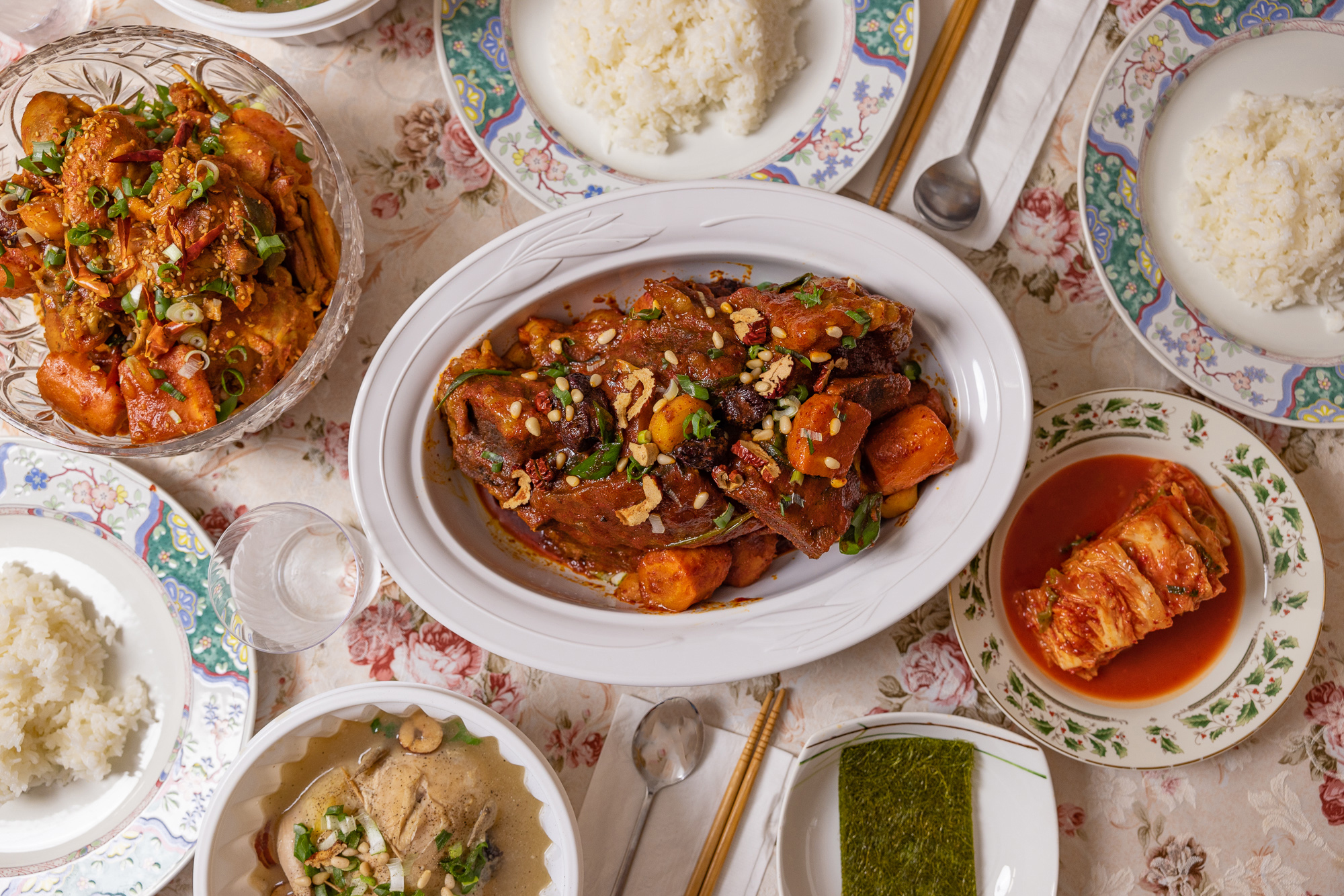 A group of plates and bowls on a flower tablecloth, with red stewed meats, a whole chicken in soup, rice, and kimchi.