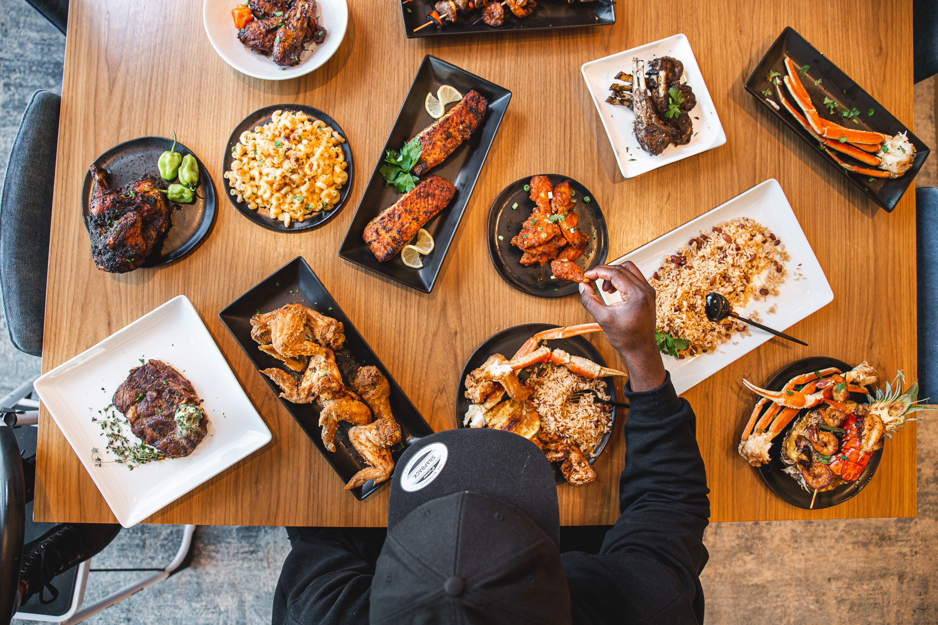 An overhead shot of Mervil sitting at a table with a spread of a dozen dishes of food in front of him. He reaches for a chicken wing with his right hand.